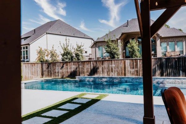 Backyard pool with patio, wooden fence, and suburban houses under a blue sky