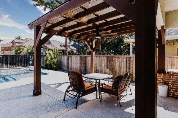 Covered patio seating beside a backyard pool, with dark wood pergola and two chairs at a small table.