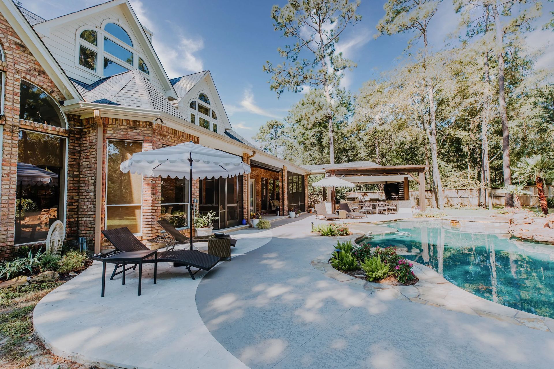 Backyard pool patio with lounge chairs, umbrella, and brick house under trees