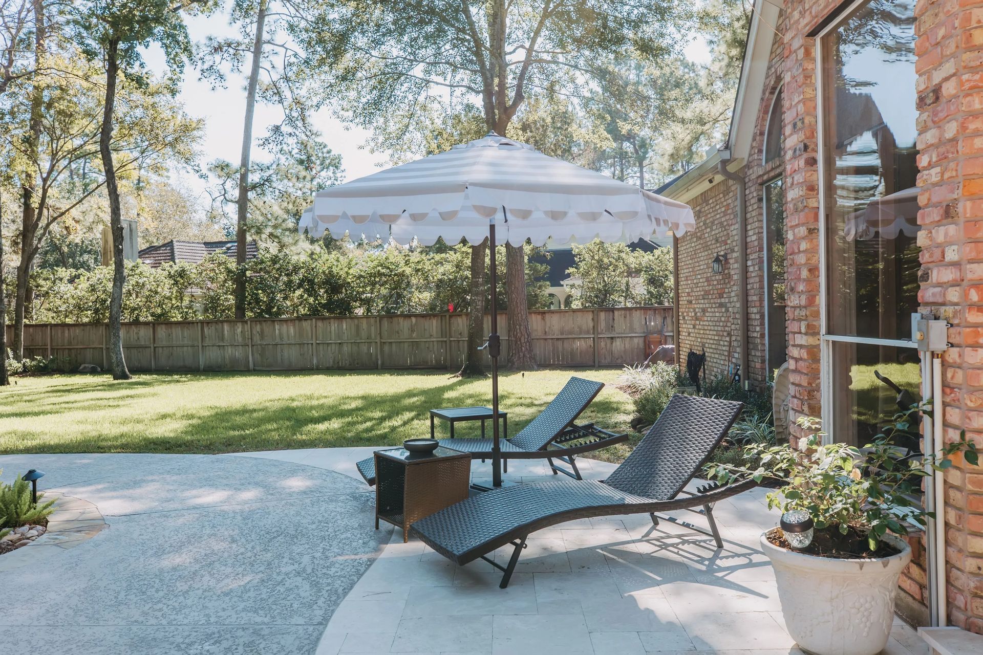 Patio with two lounge chairs, a white umbrella, potted plants, and a brick house beside a lawn and trees