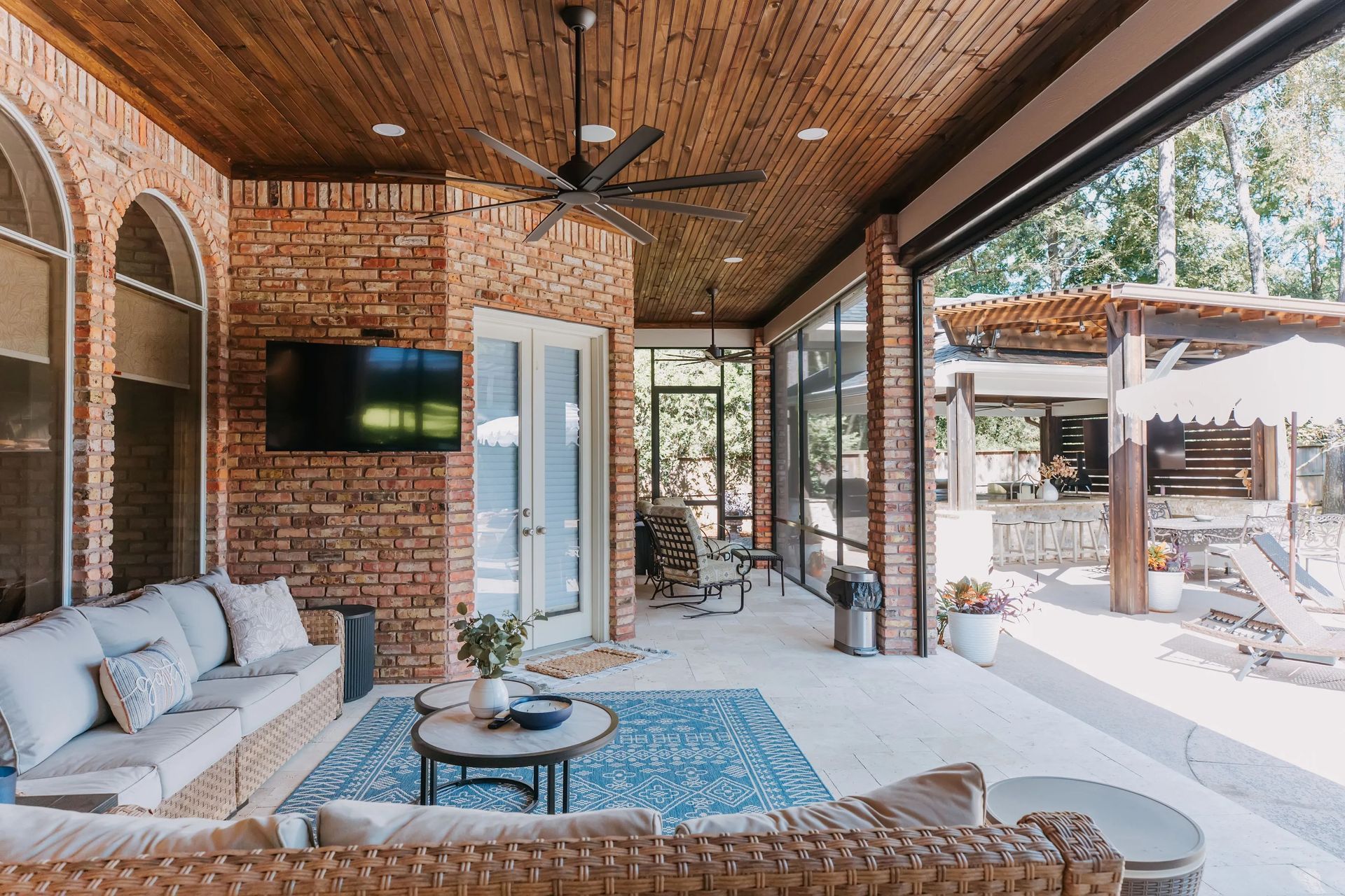 Covered brick patio with wicker seating, wall-mounted TV, and bright outdoor dining area