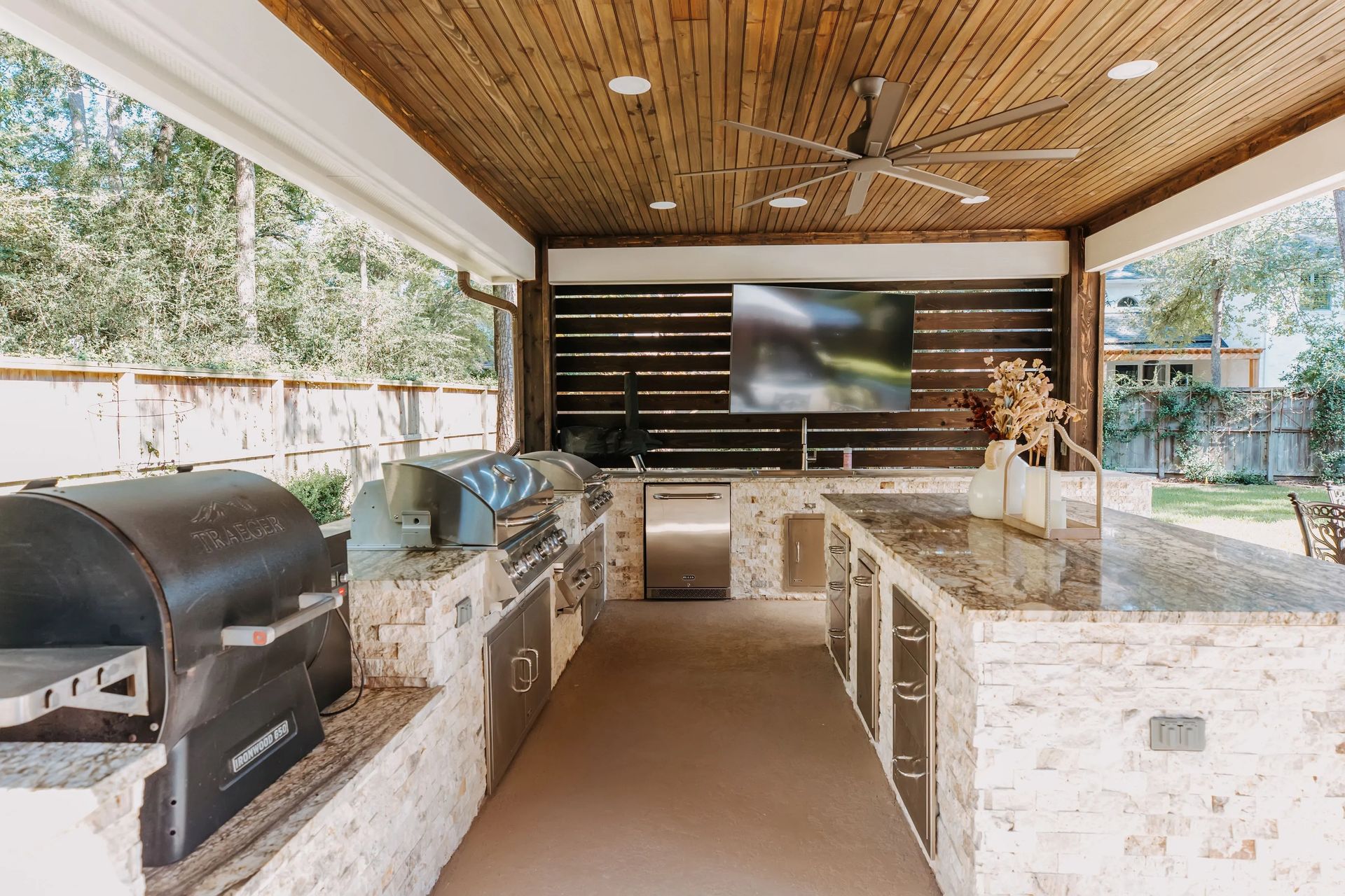 Outdoor covered patio with stone kitchen, built-in grill, TV, and wood-slat ceiling
