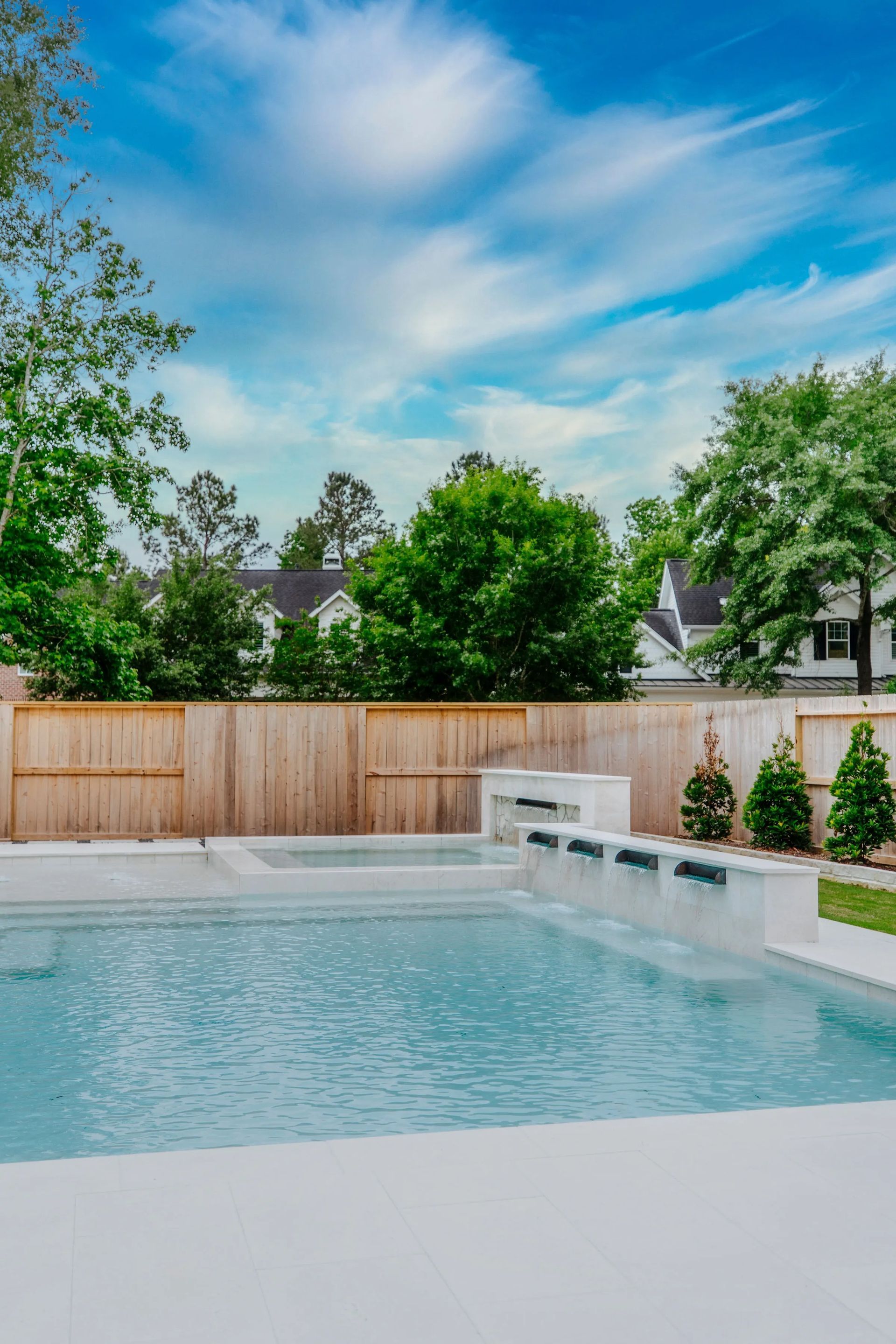 Backyard swimming pool with blue water, white deck, wooden fence, and green trees under a bright sky