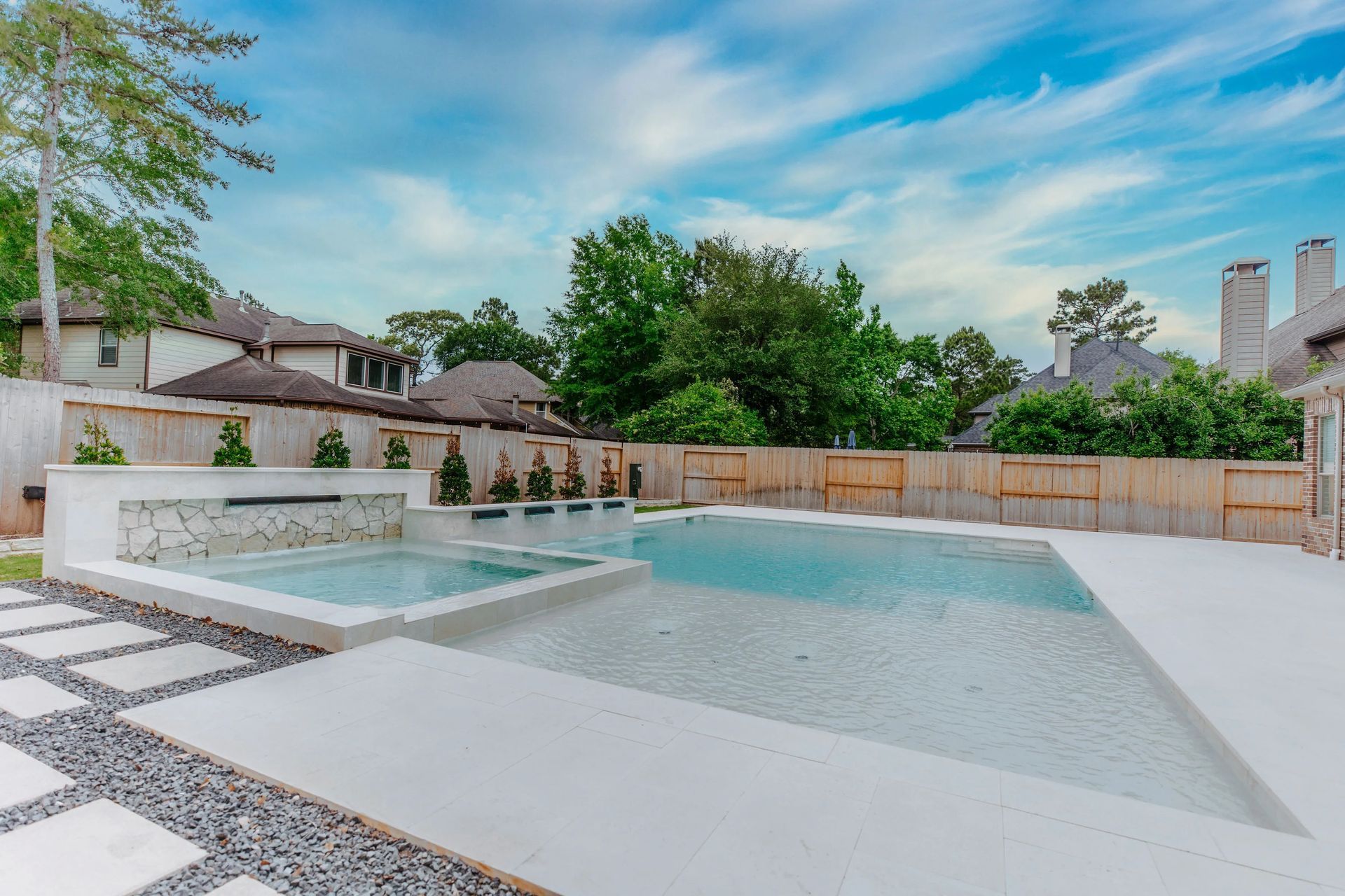 Modern backyard pool with a shallow ledge, white deck, gravel border, and trees under a blue sky
