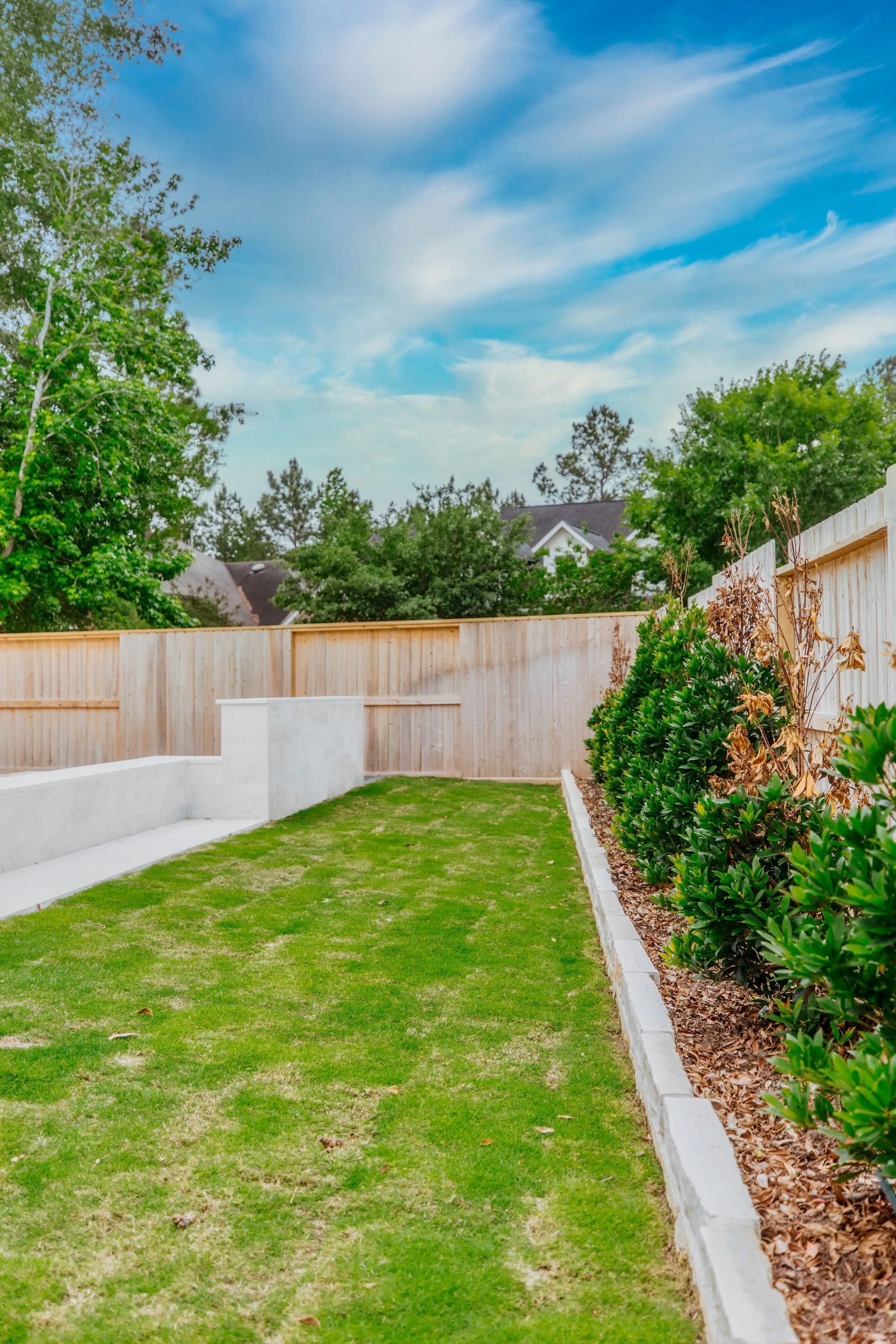 Narrow backyard lawn with white retaining walls, wooden fence, shrubs, and a blue sky