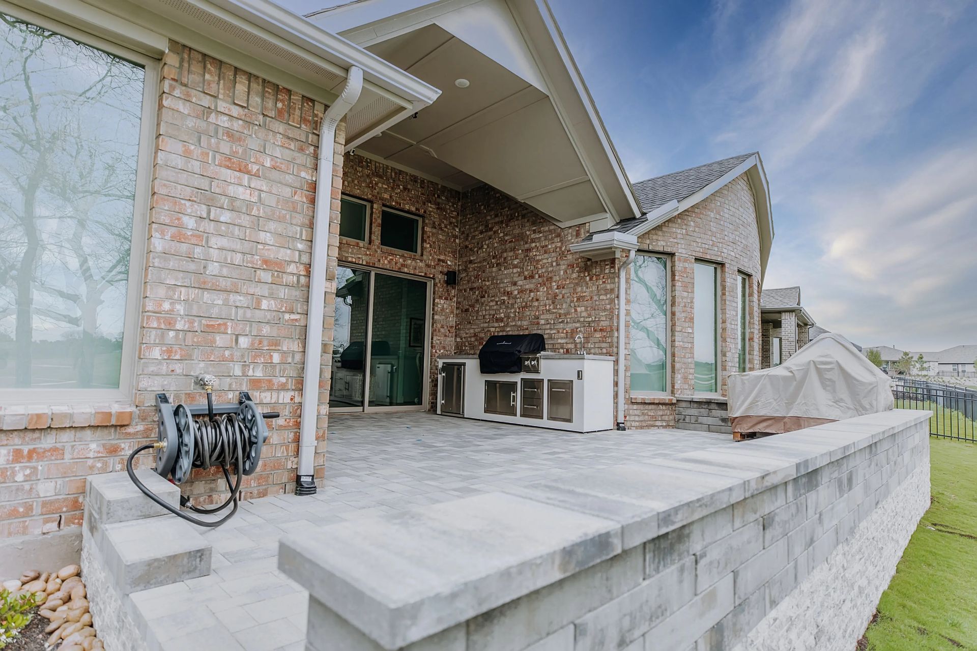 Brick house patio with concrete steps, outdoor sink, grill, and large windows overlooking a lawn