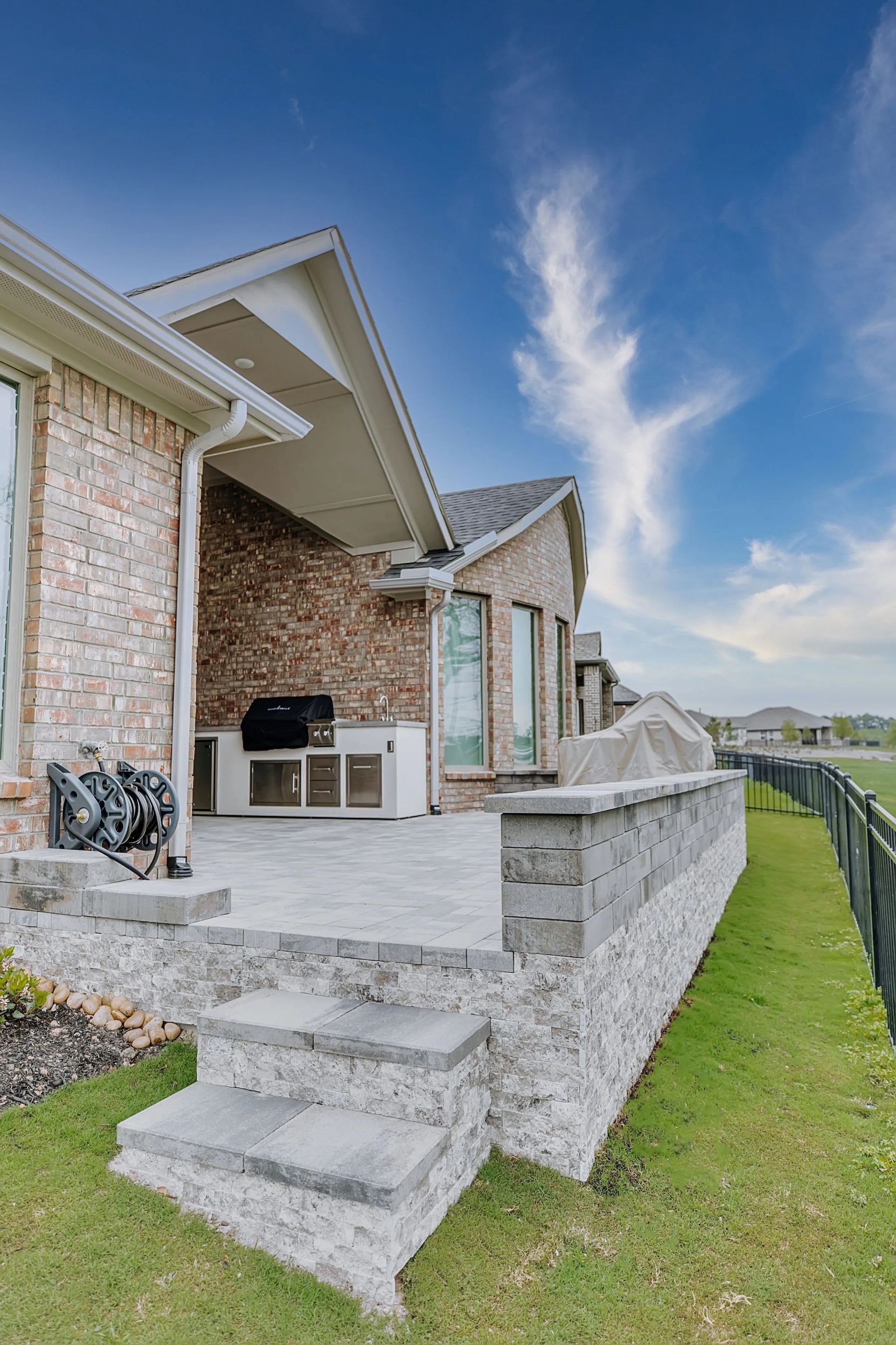 Brick house patio with steps, outdoor grill, and a stone retaining wall beside a green lawn.