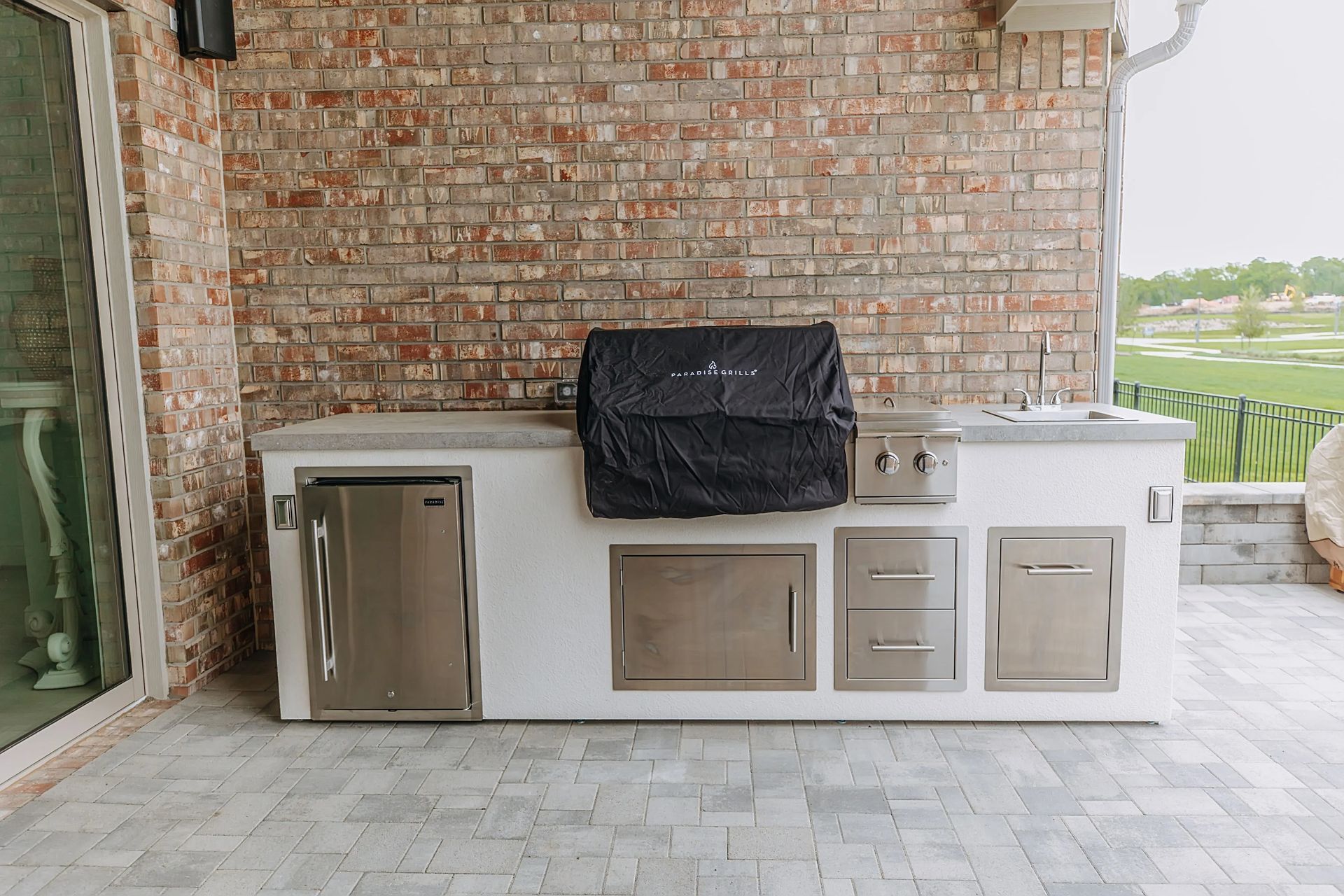 Covered outdoor patio with a stainless grill, mini fridge, and white storage cabinets against a brick wall