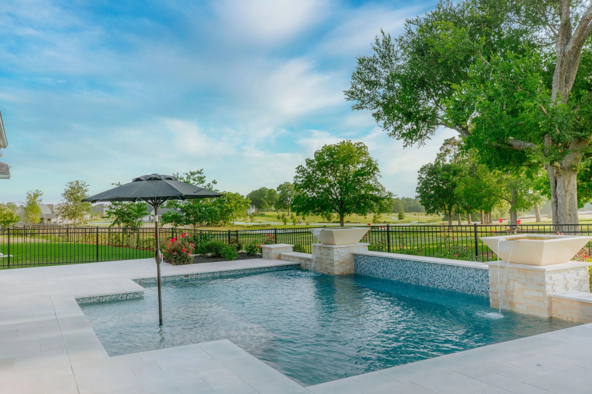 Backyard pool with umbrella, stone patio, and trees under a blue sky