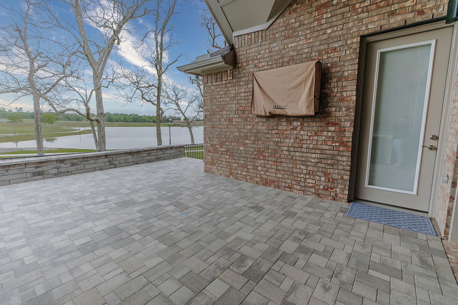 Brick patio beside a house with a glass door, covered window, and view of leafless trees and water beyond