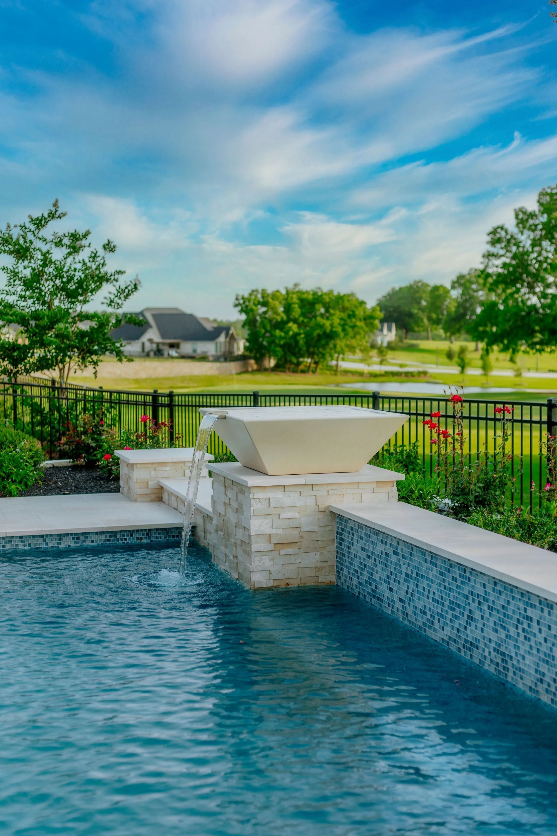 Backyard pool with a stone waterfall, black fence, green lawn, and blue sky beyond.