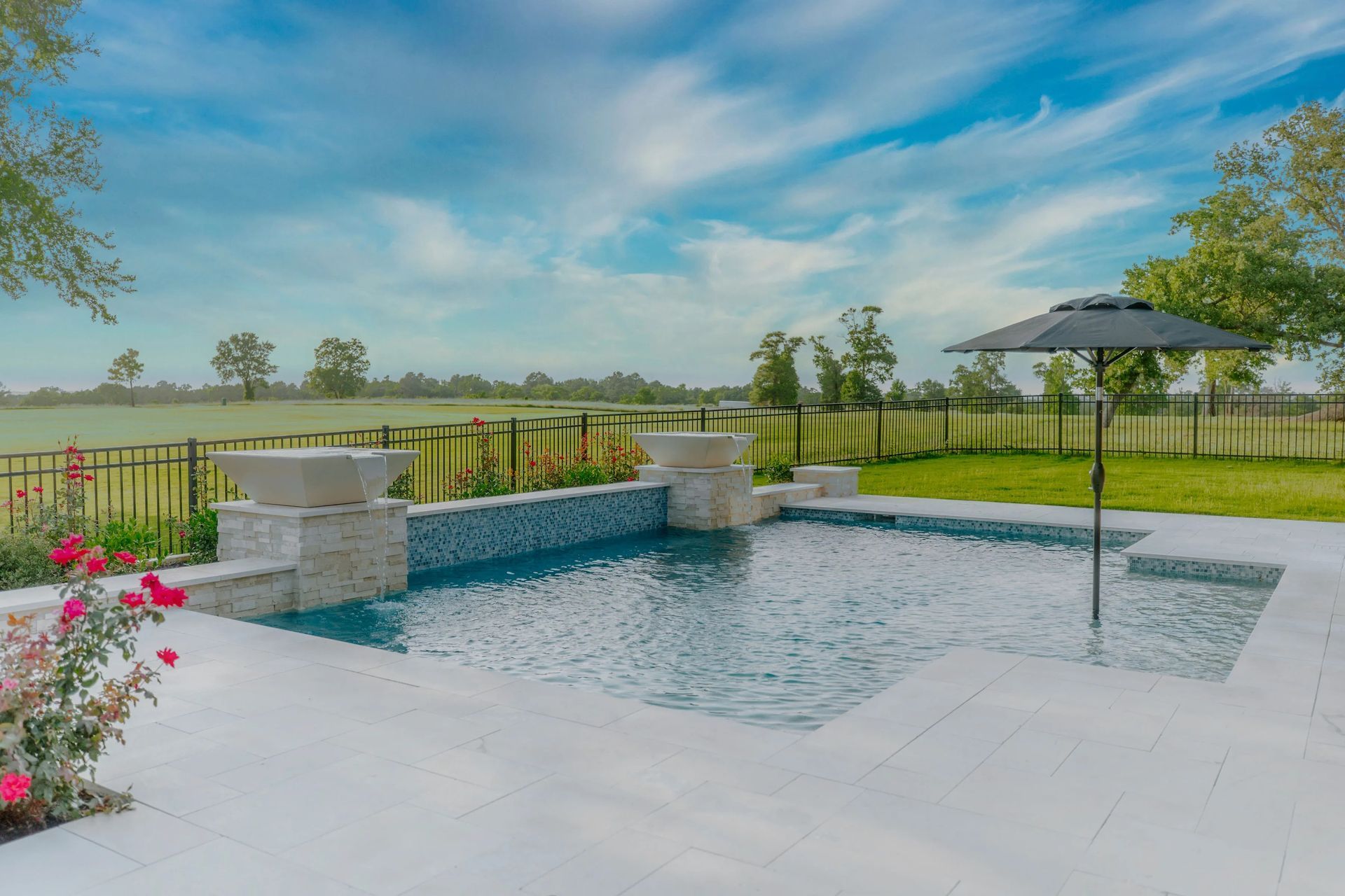 Backyard swimming pool with stone water feature, patio, and umbrella under a blue sky