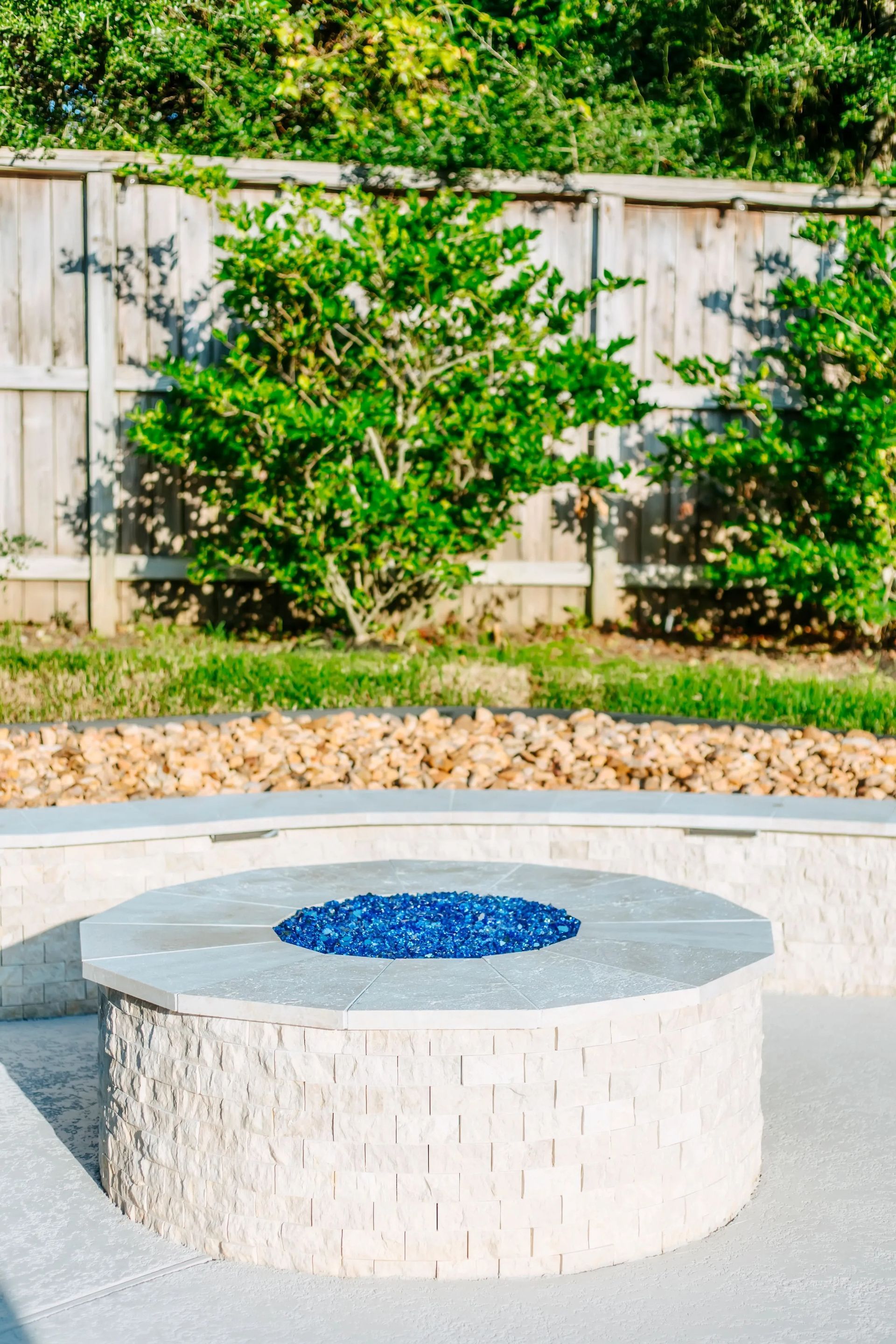 Round white stone fire pit with blue glass center in a landscaped backyard.