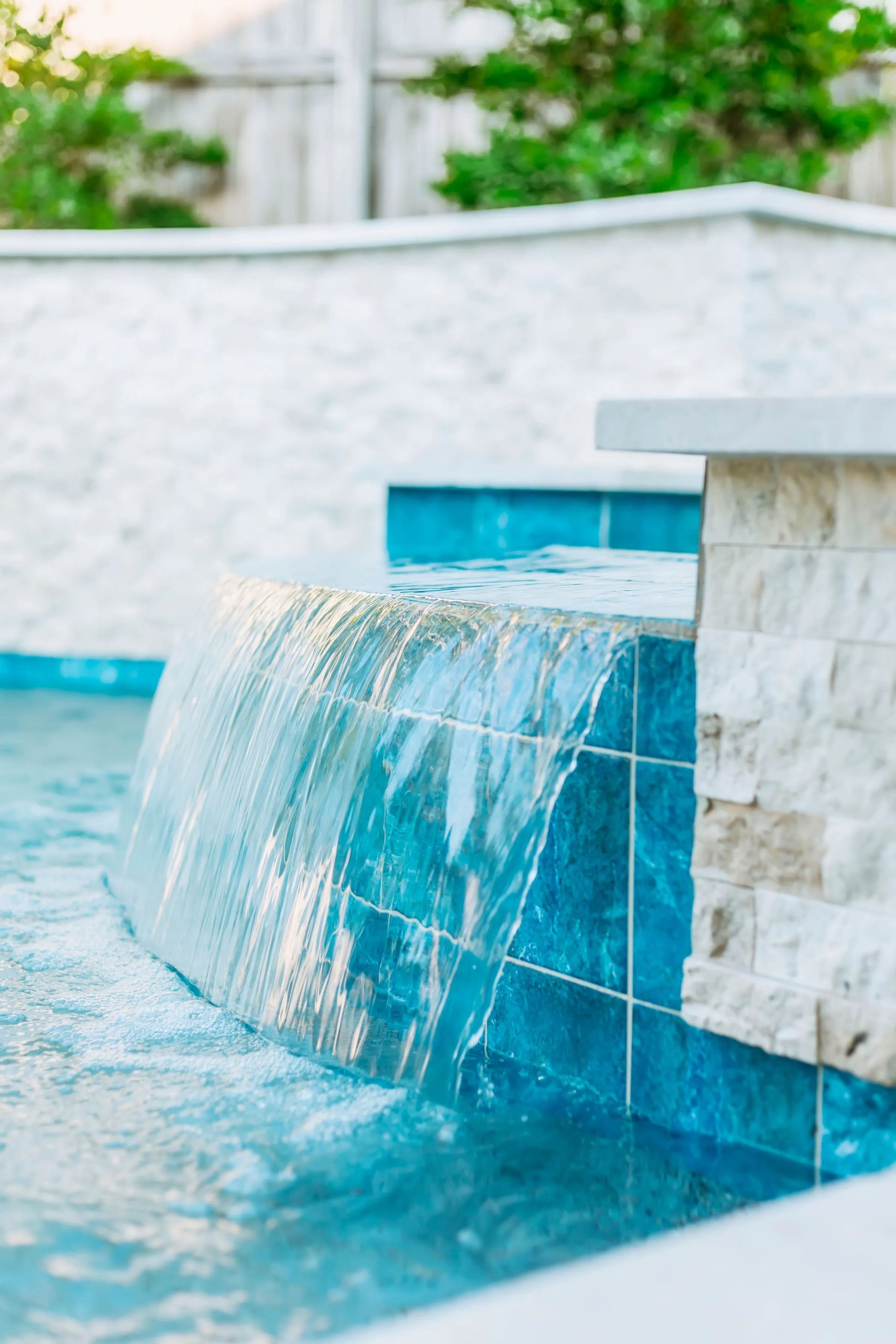 Blue-tiled pool waterfall spilling into clear water beside a white stone ledge
