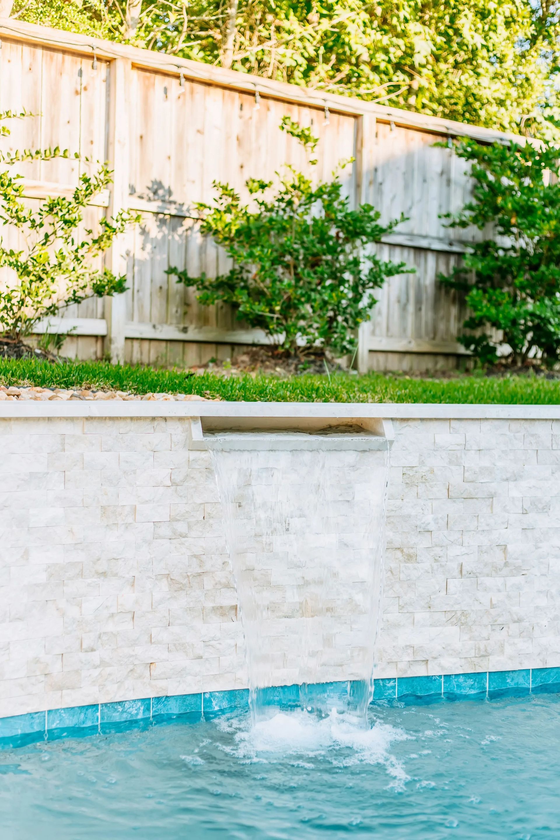 Backyard pool with a white stone wall, wooden fence, and greenery above the water.