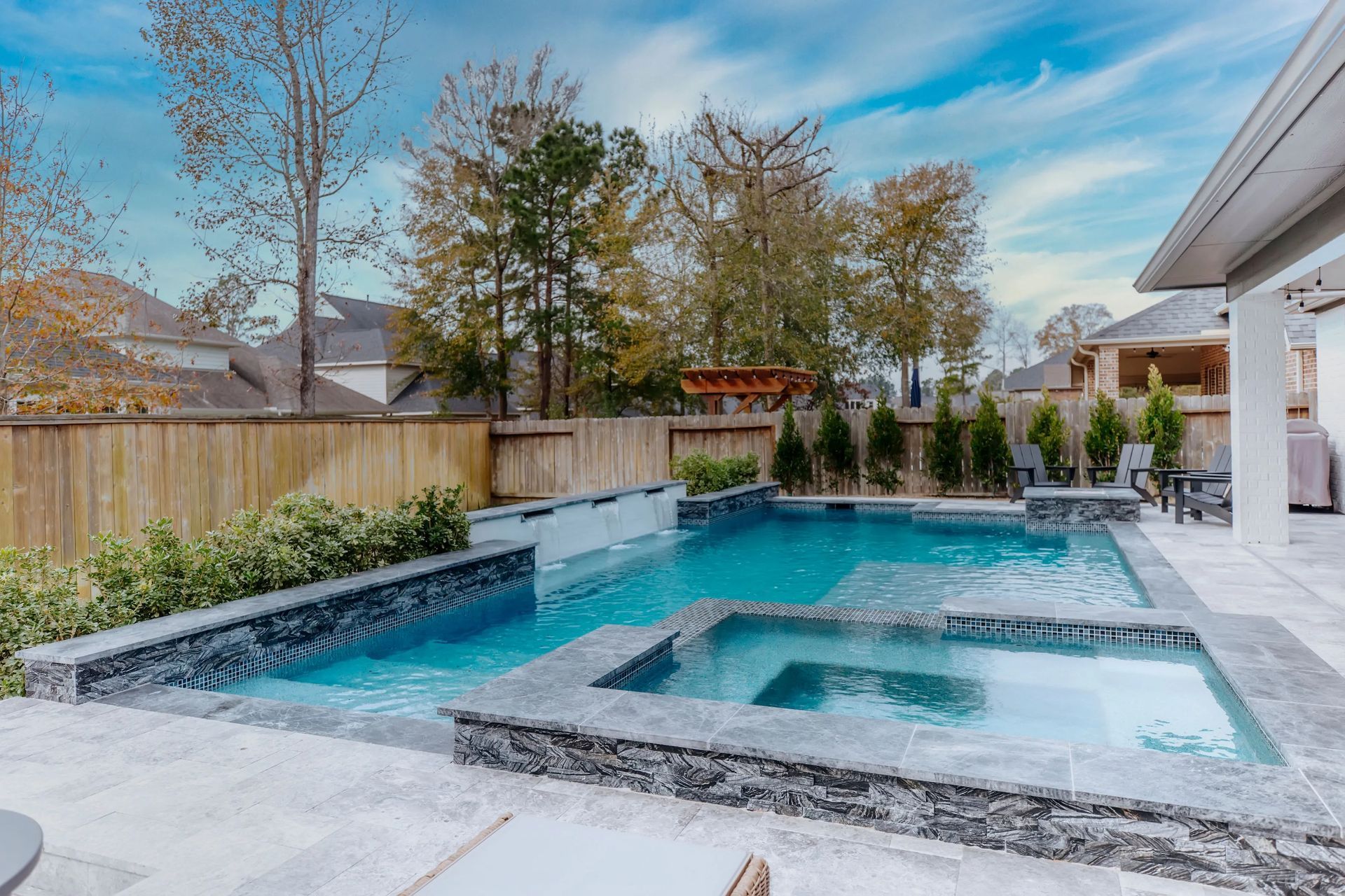 Backyard patio with a turquoise swimming pool, spa, stone decking, and wood fence under a blue sky