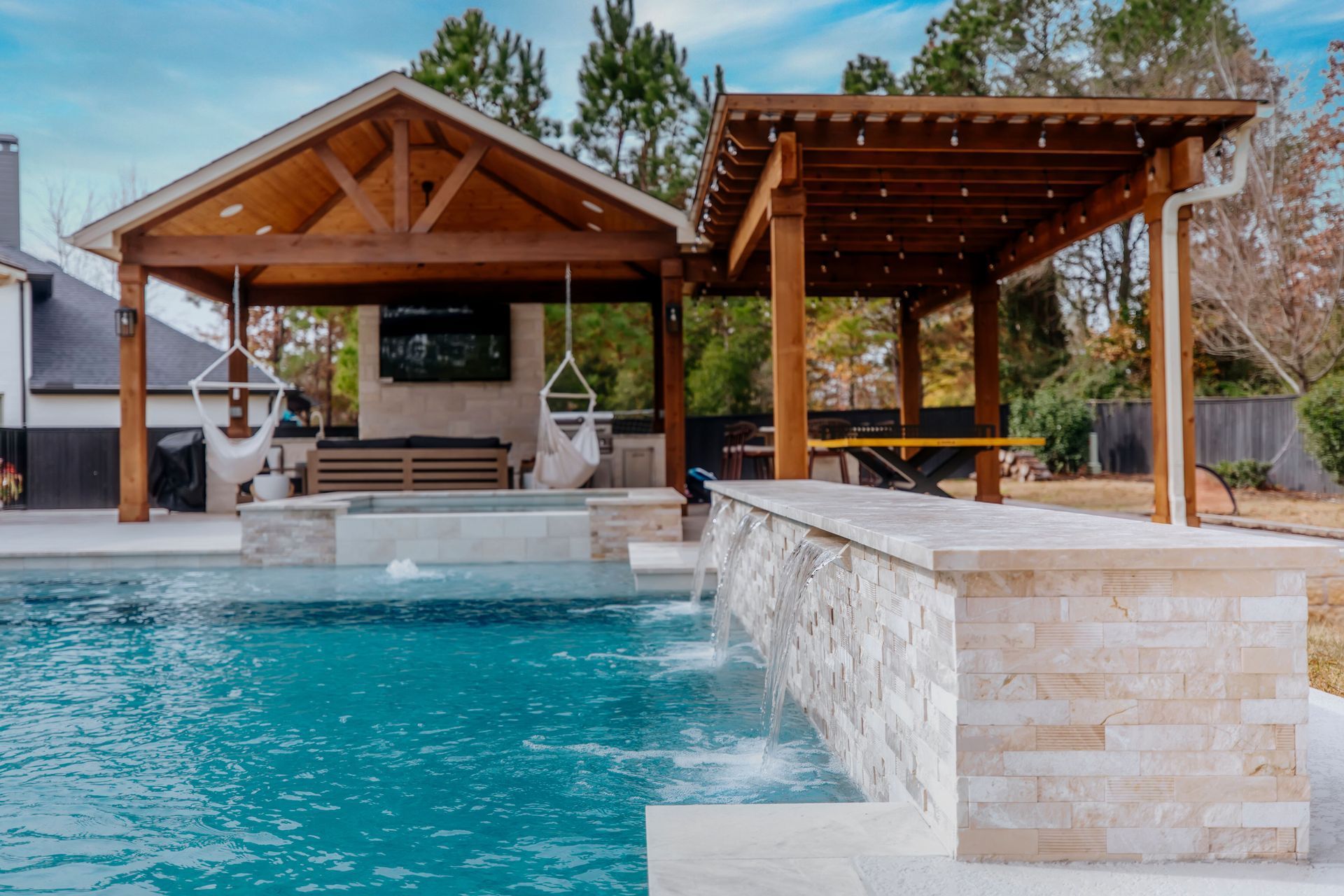 Backyard pool with a wood pavilion, pergola seating area, and stone water feature