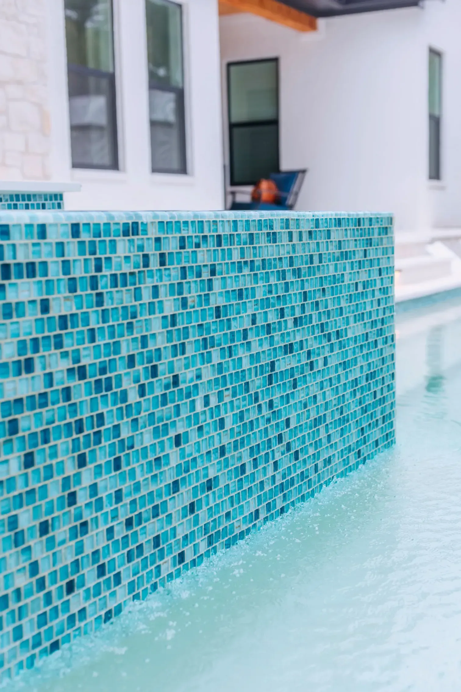 Turquoise-tiled pool edge beside a white house with a small potted plant on the patio