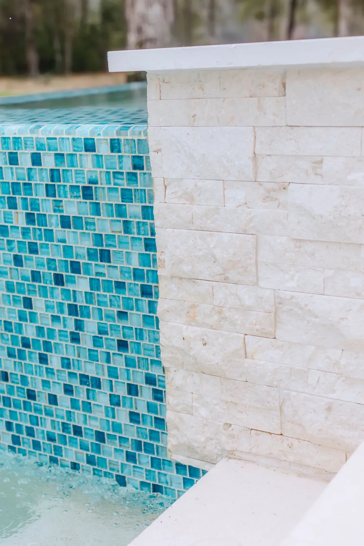 Blue mosaic pool wall beside a beige stone ledge with water at the bottom