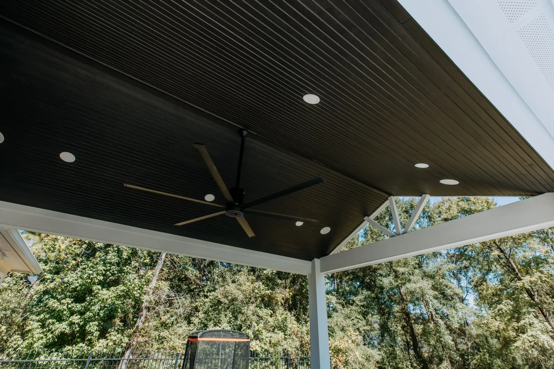 Outdoor patio ceiling with a black fan, recessed lights, and trees visible beyond the white columns
