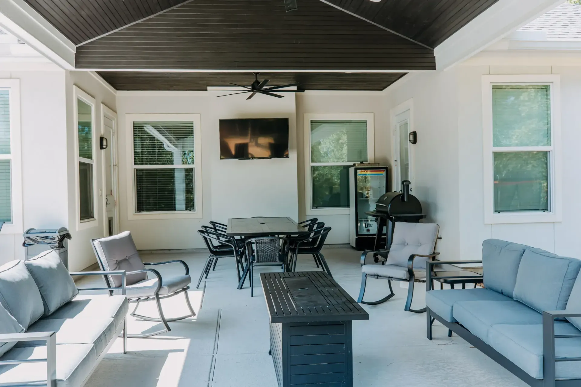 Covered patio with blue-gray seating, dining table, and wall-mounted TV under a dark ceiling