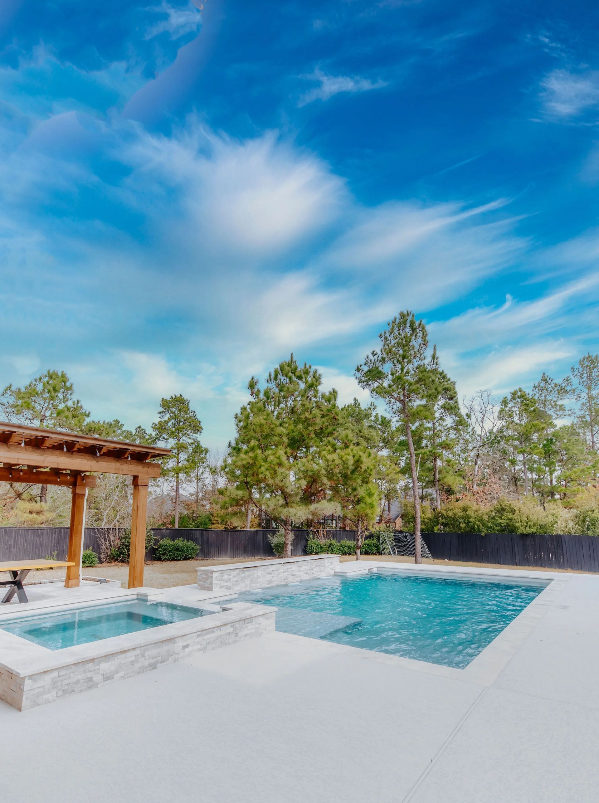 Backyard pool and hot tub with white patio, wooden pergola, and trees under a blue sky