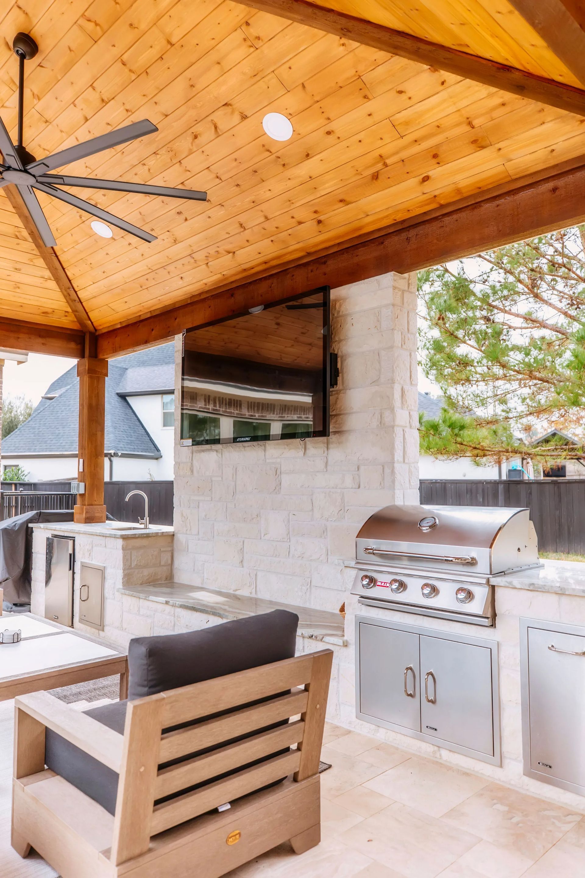 Covered outdoor patio with wood ceiling, ceiling fan, grill, and seating area by a stone wall