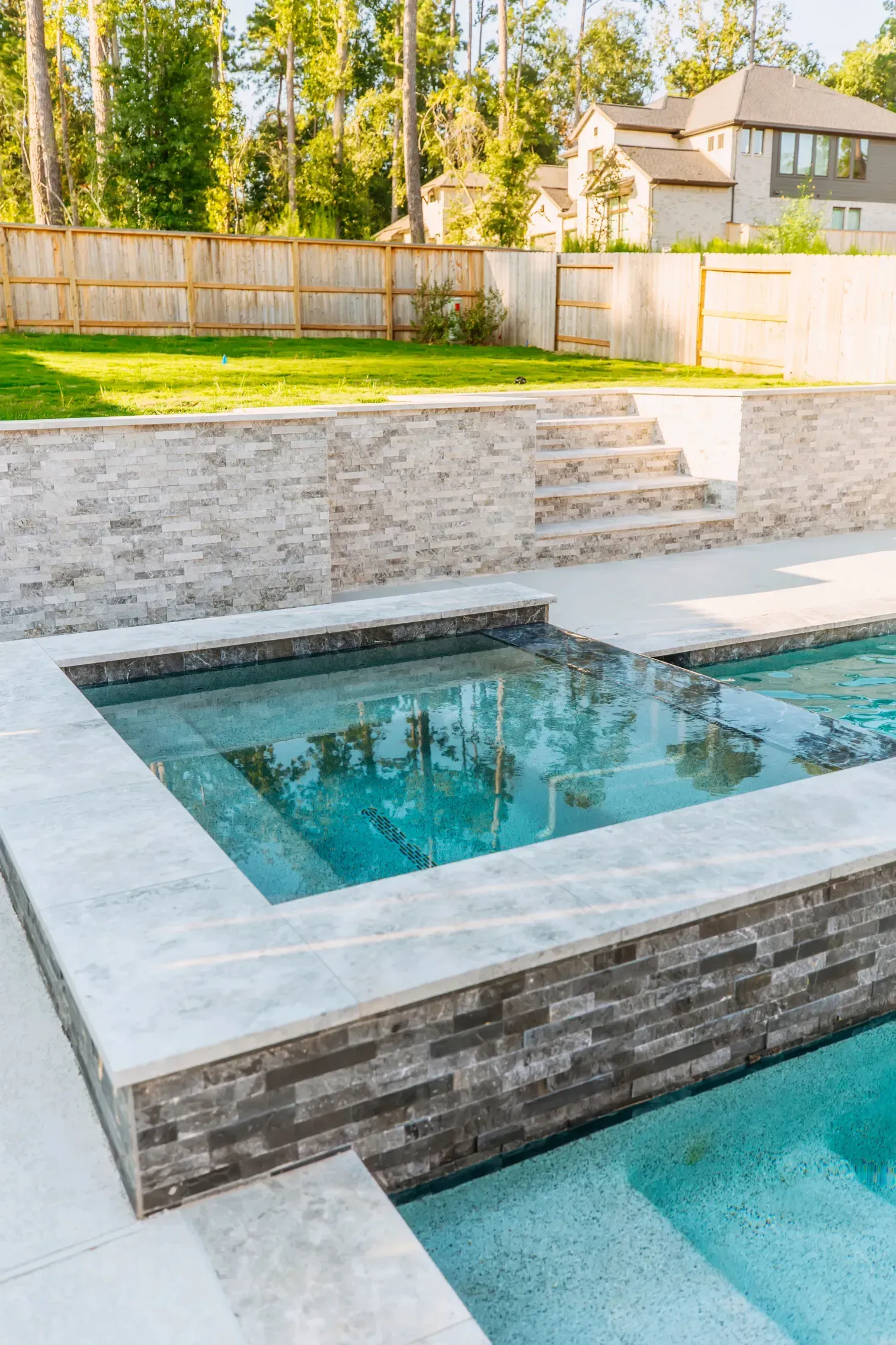 Backyard with modern tiered pool and white stone coping beside a fenced lawn