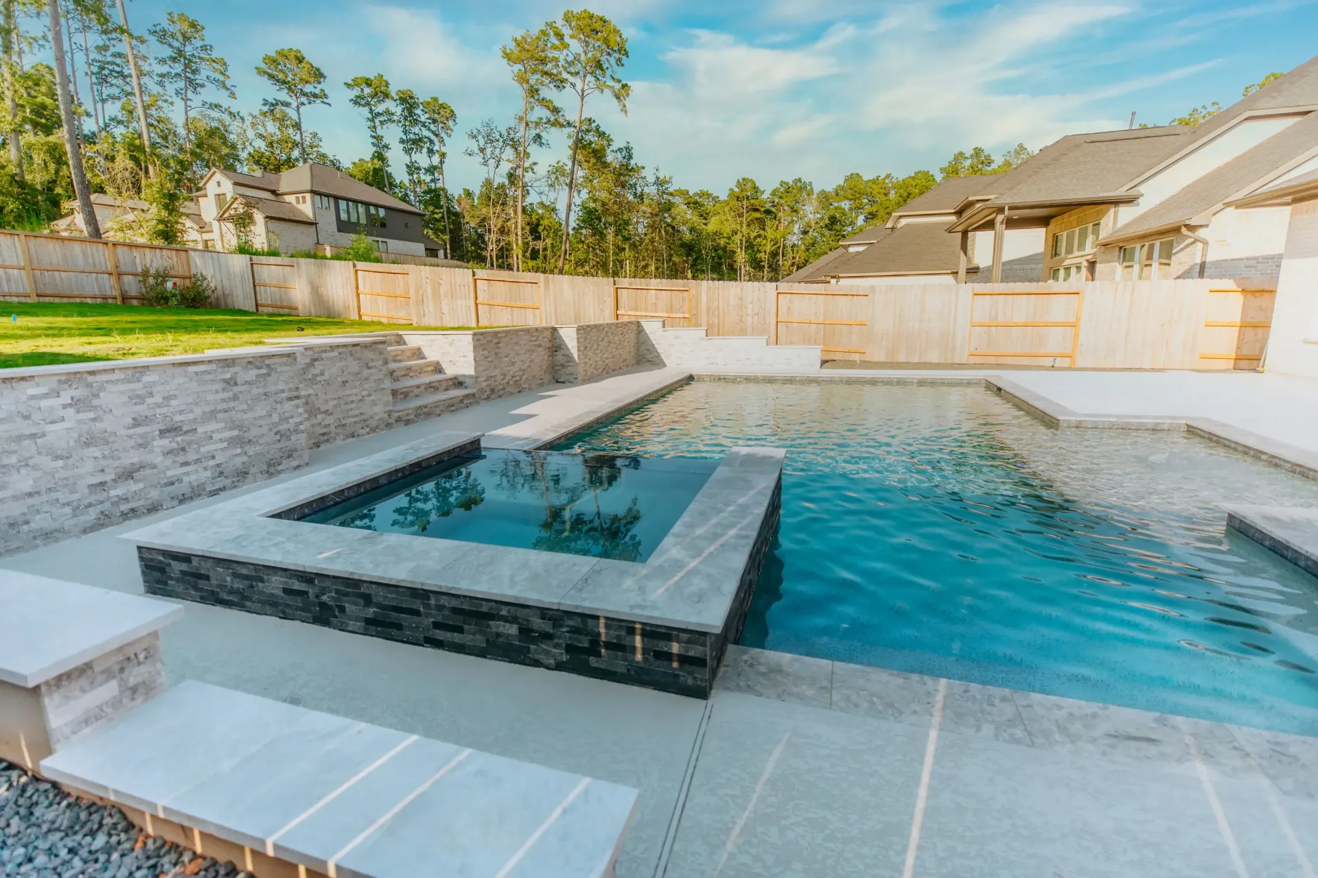 Modern backyard patio with a turquoise pool and raised square spa beside stone seating and a wooden fence