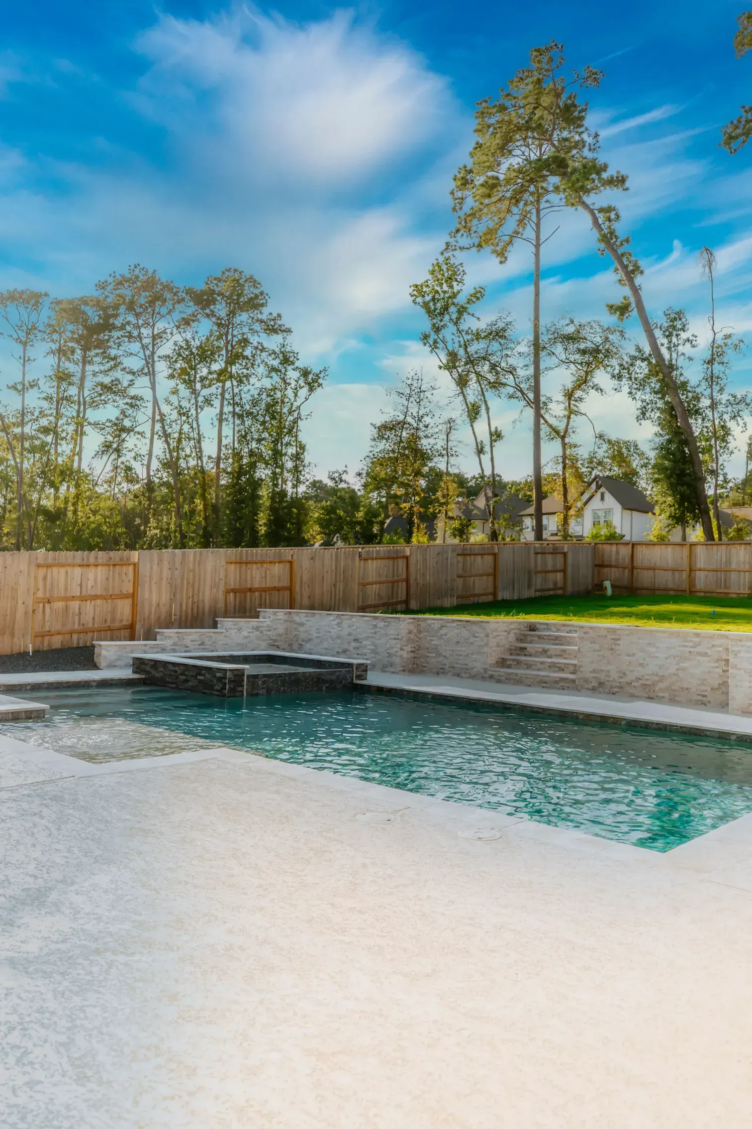 Modern backyard pool with stone deck, wooden fence, and tall trees under a blue sky