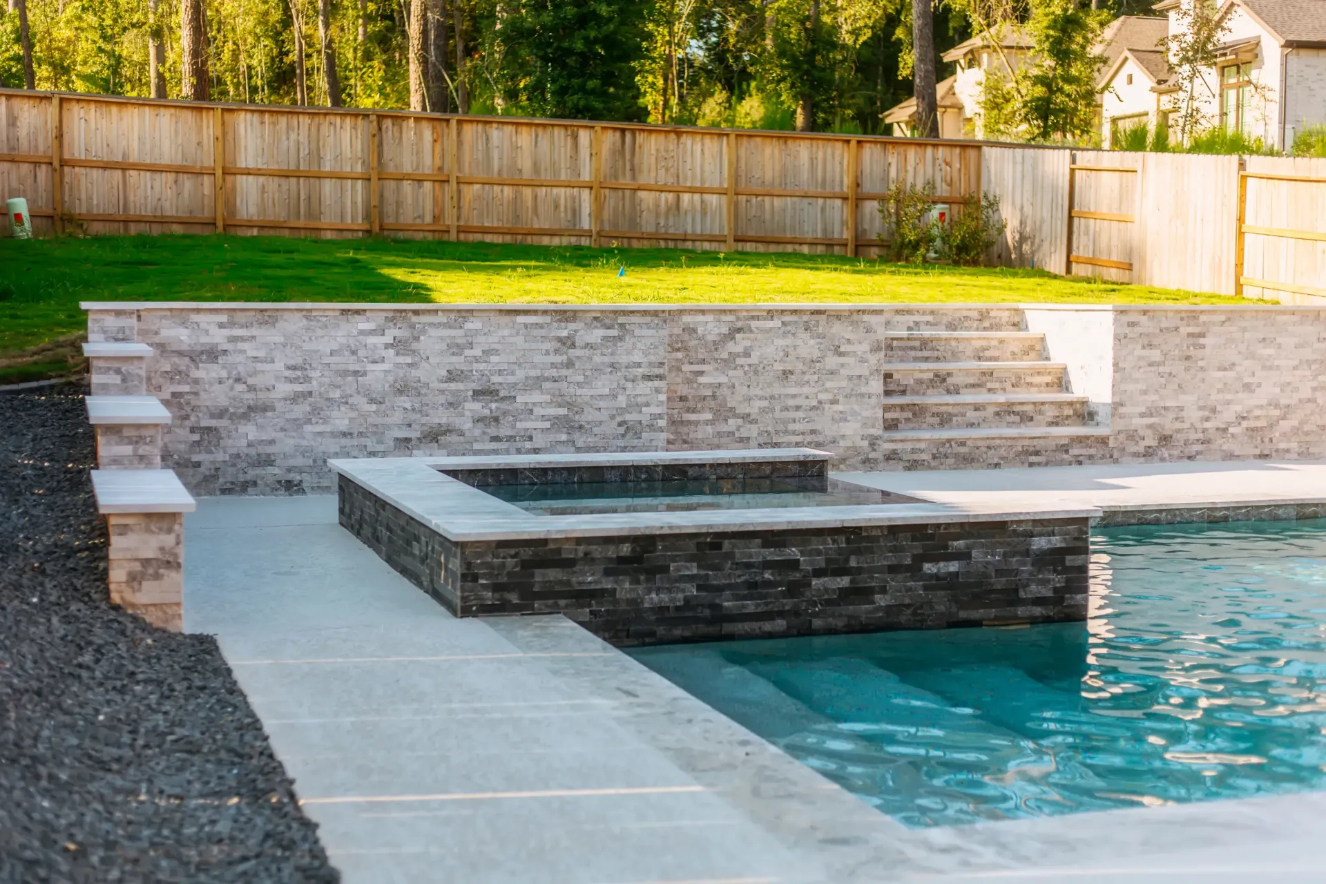 Modern backyard pool with stone patio, raised spa, and wooden fence backdrop