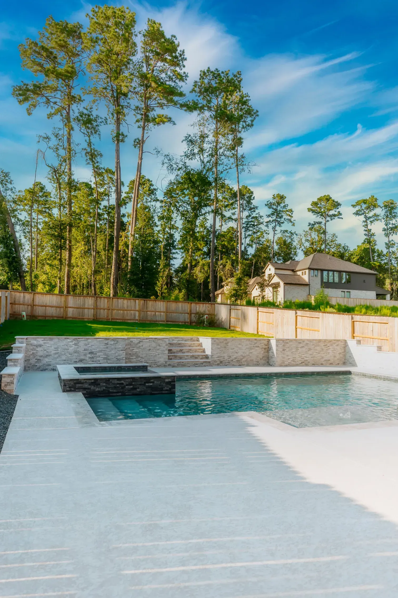 Modern backyard with a rectangular pool, stone water feature, and tall trees behind a fence