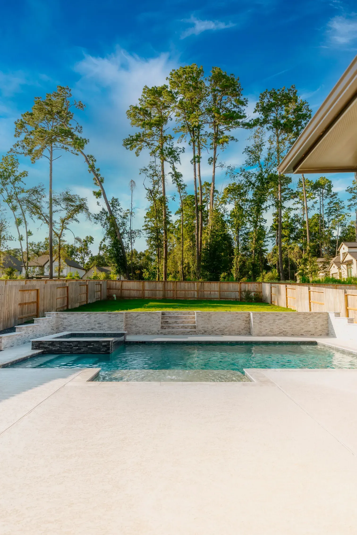 Modern backyard pool with a turquoise water feature, white patio, and tall trees under a blue sky