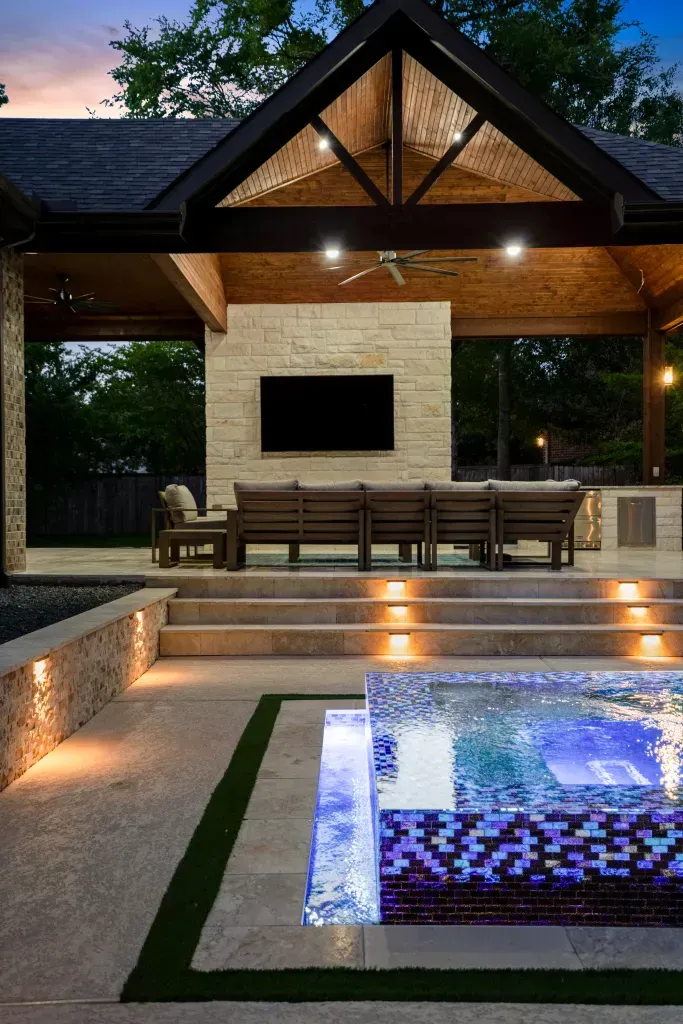 Modern backyard patio with lit steps, a long table, and a glowing pool beside a covered pavilion at dusk