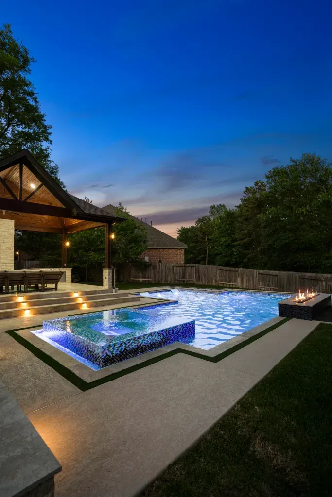 Modern backyard pool at dusk with blue LED lighting and a covered patio nearby