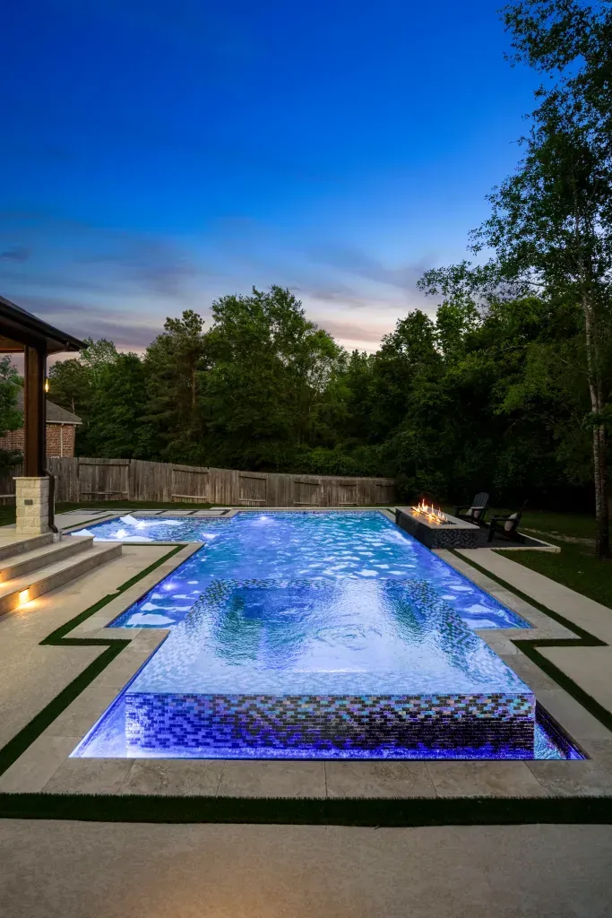 Backyard pool glowing blue at dusk, surrounded by trees and a stone patio