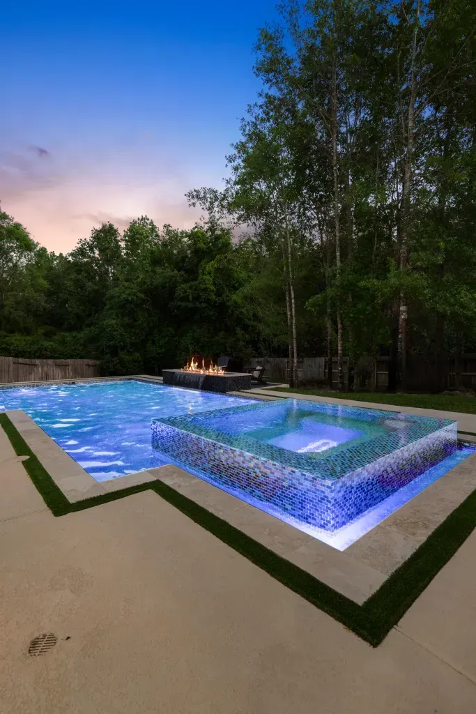 Backyard pool with glowing blue water, surrounding deck, and trees at dusk