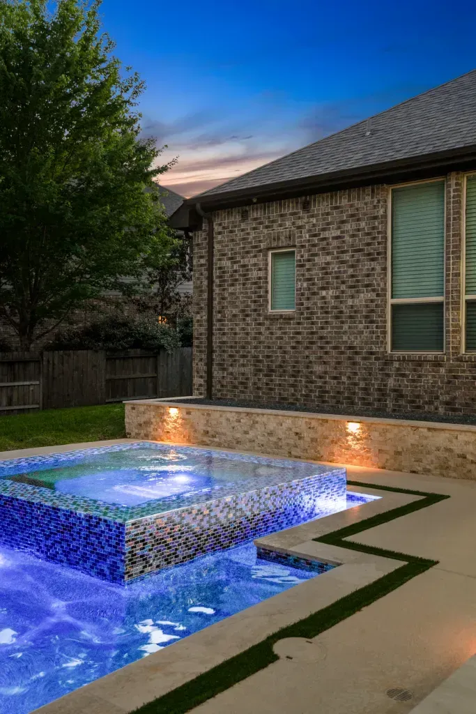 Backyard pool with glowing blue water beside a brick house at dusk