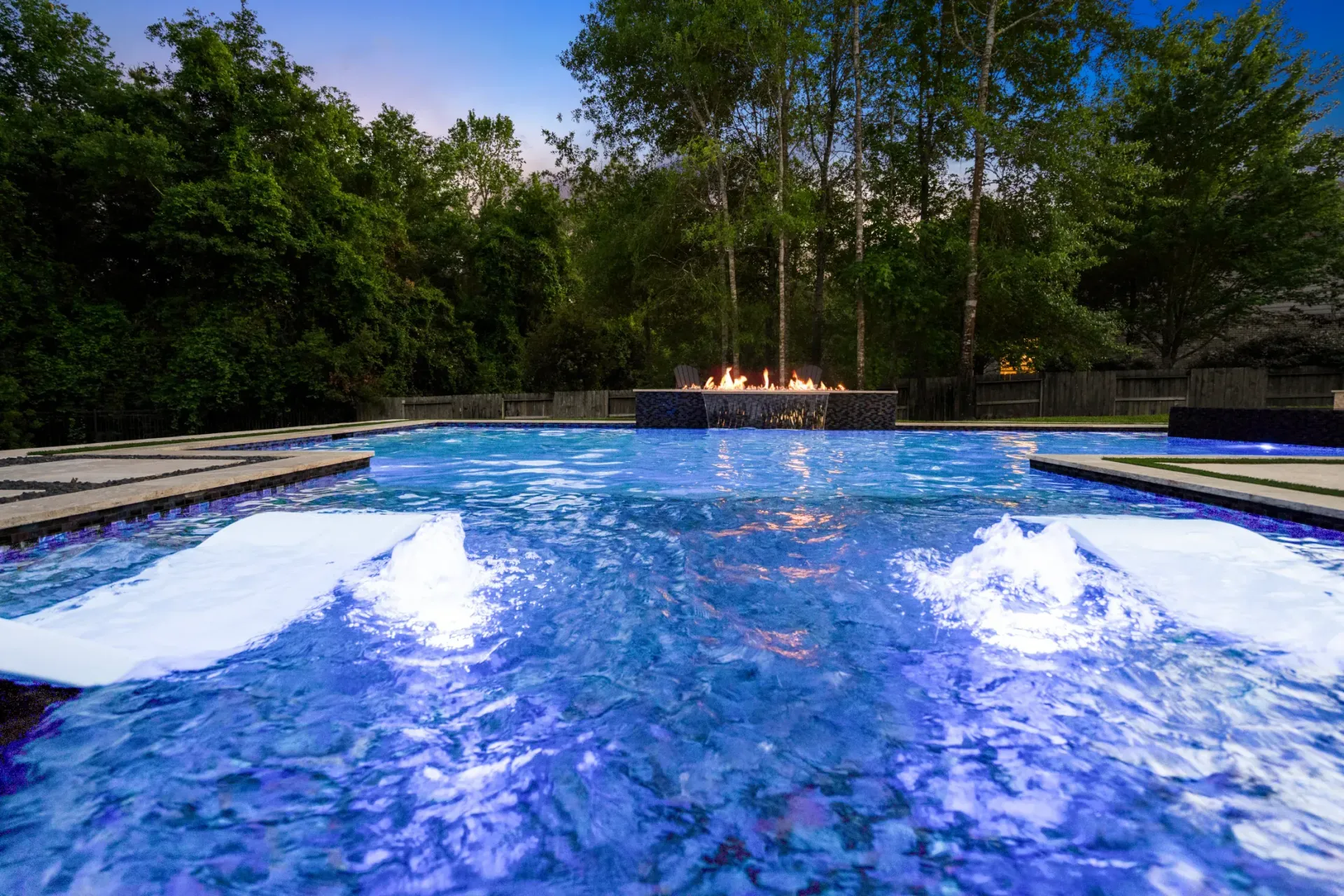 Blue pool with bubbling water jets, stone edge, and trees in the background at dusk
