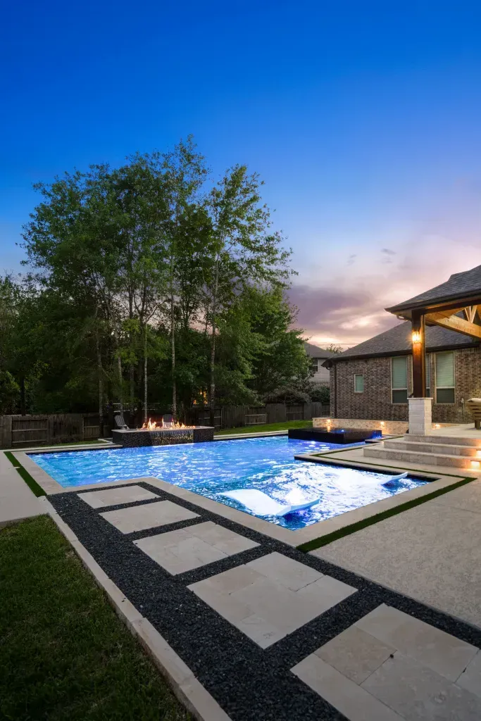 Modern backyard pool at dusk with blue-lit water, stone patio, trees, and a covered seating area