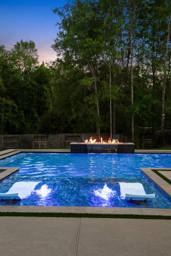 Lit backyard pool at dusk with blue lights and a fire feature beyond the water