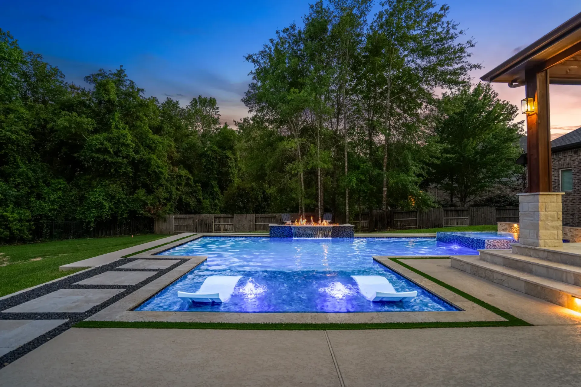 Lit backyard pool and spa at dusk with blue lights, stone deck, and trees surrounding it