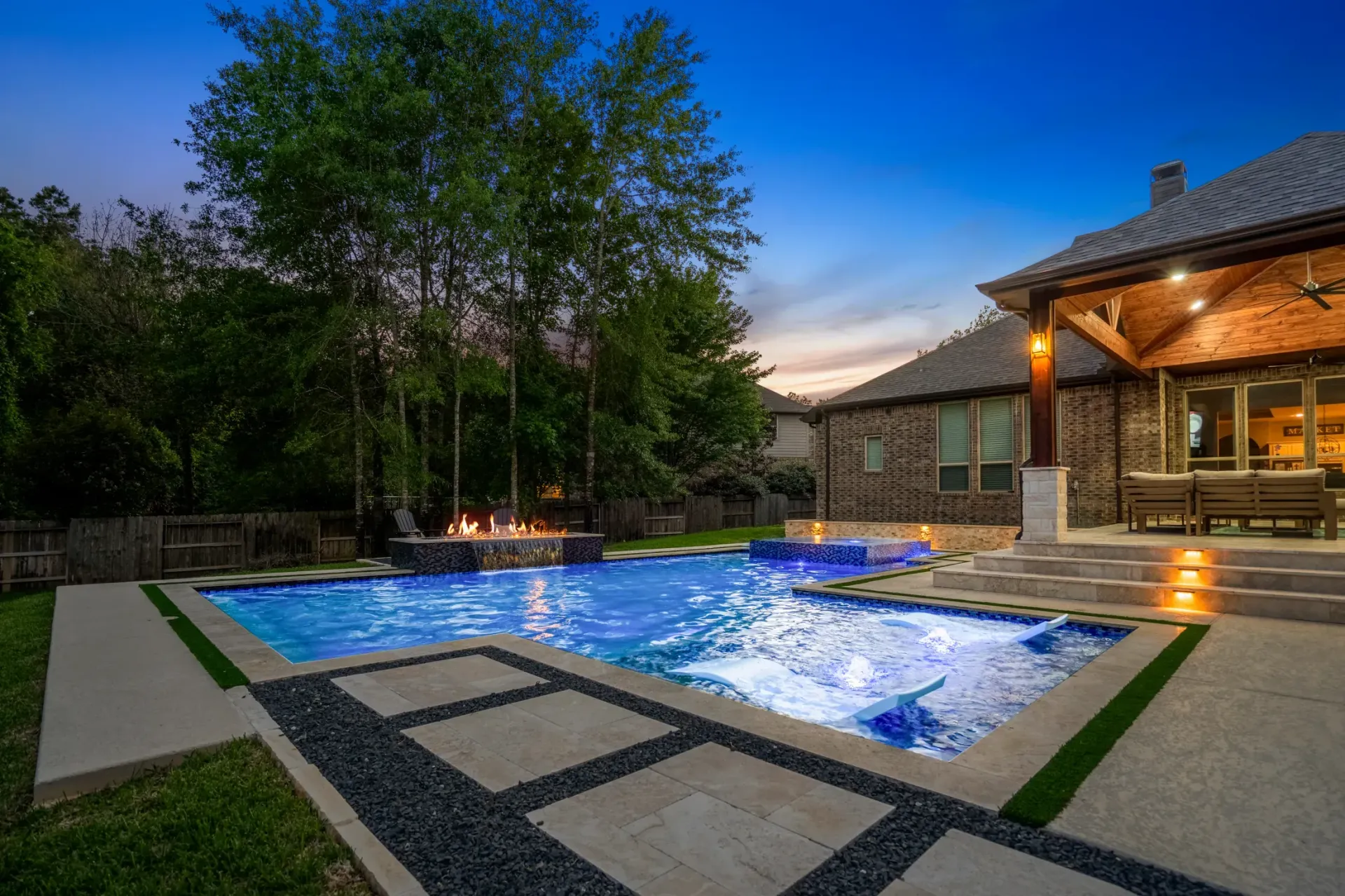 Backyard pool with glowing blue water beside a lit patio and trees at dusk