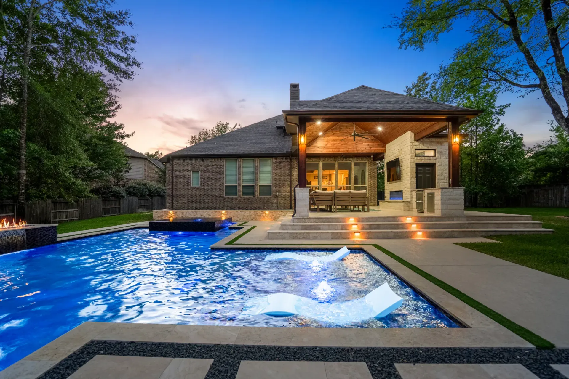 Modern brick house with lit patio and illuminated backyard pool at dusk