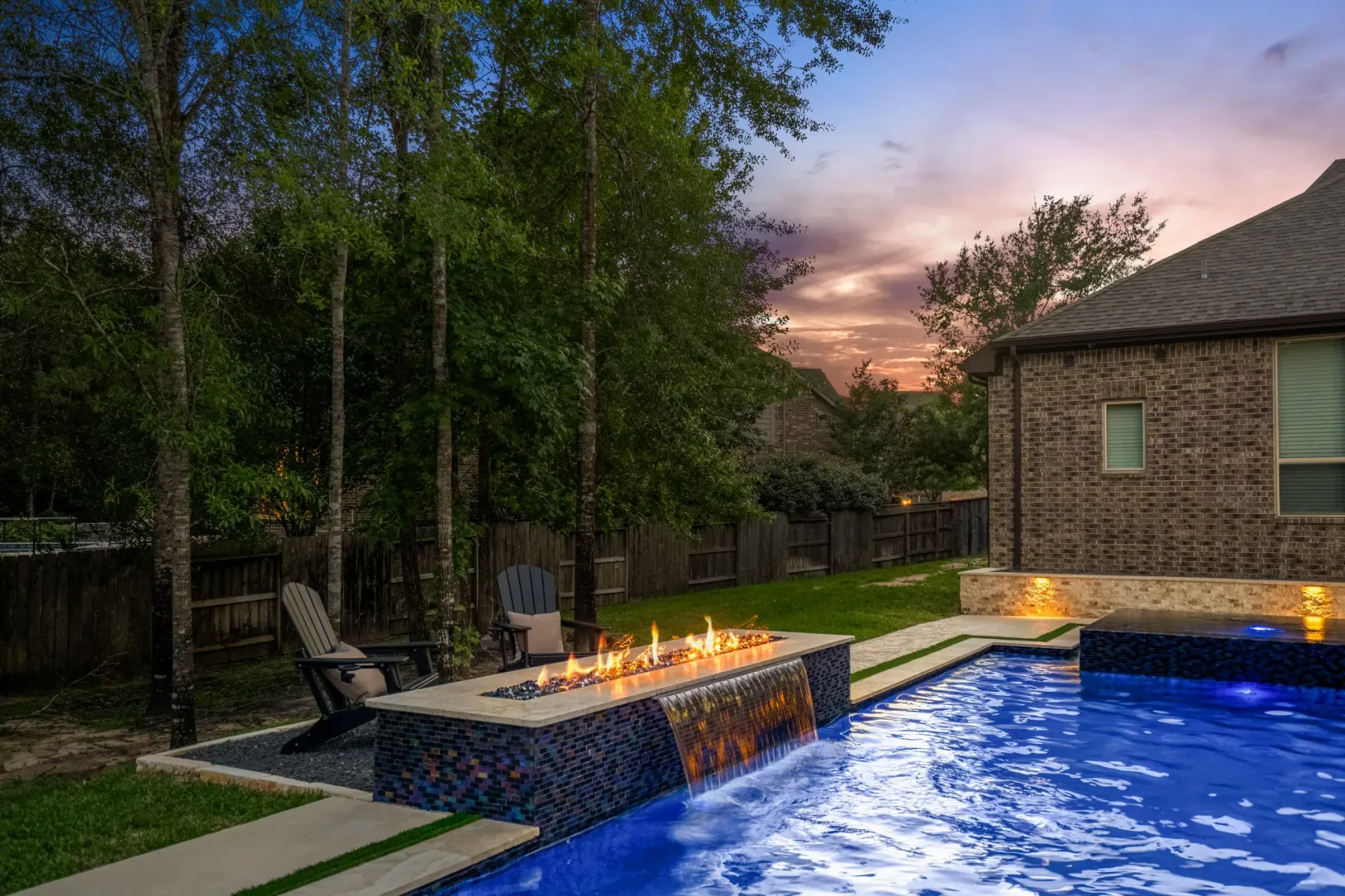 Backyard pool and spa at dusk with blue water, stone patio, and a lit fire feature beside a house