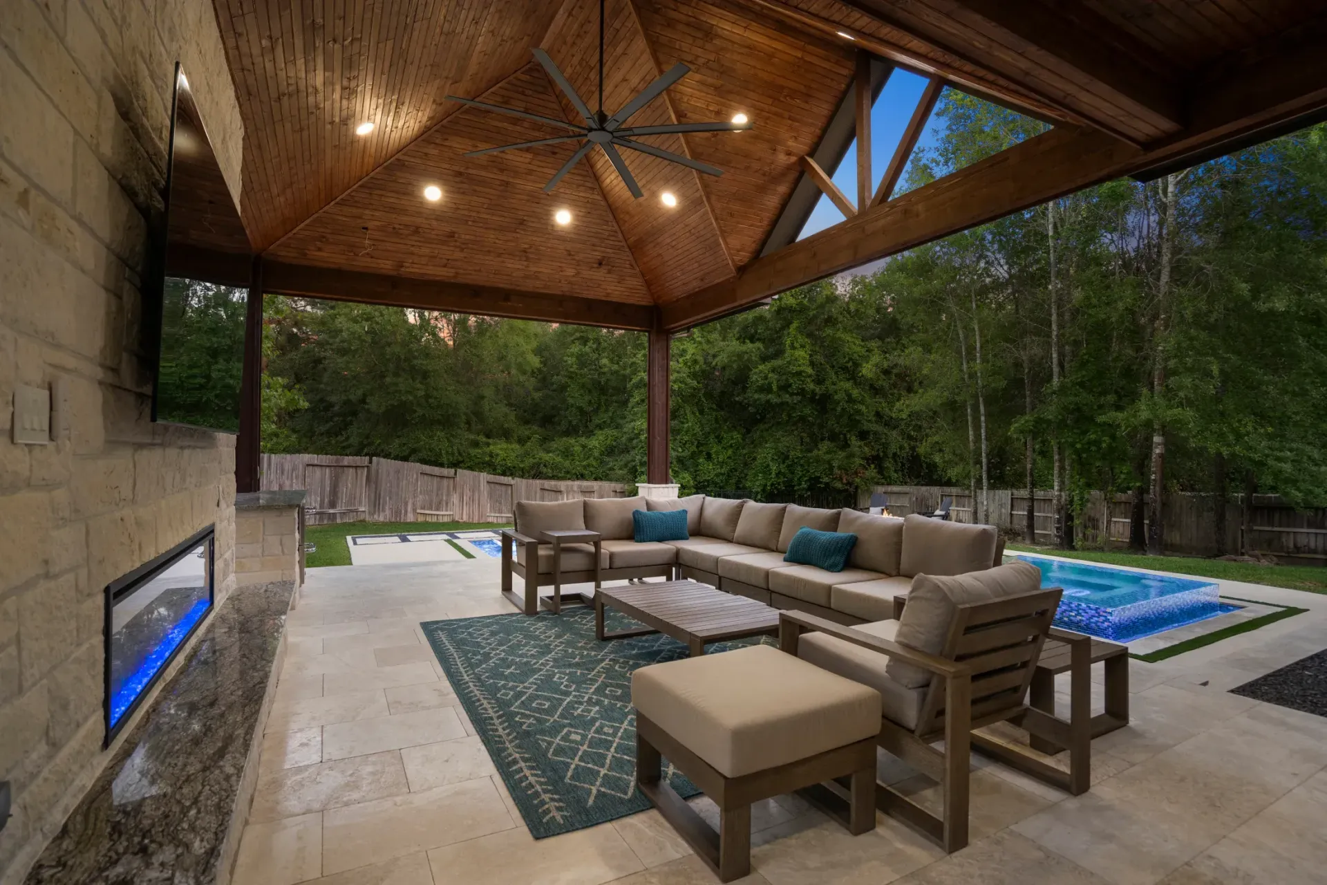 Covered outdoor patio with wicker seating, fireplace, rug, and pool beside wooded backyard.
