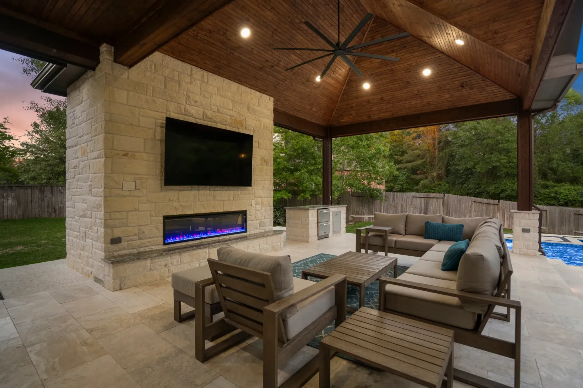 Covered stone patio with TV, blue-lit fireplace, and outdoor seating under a wooden ceiling