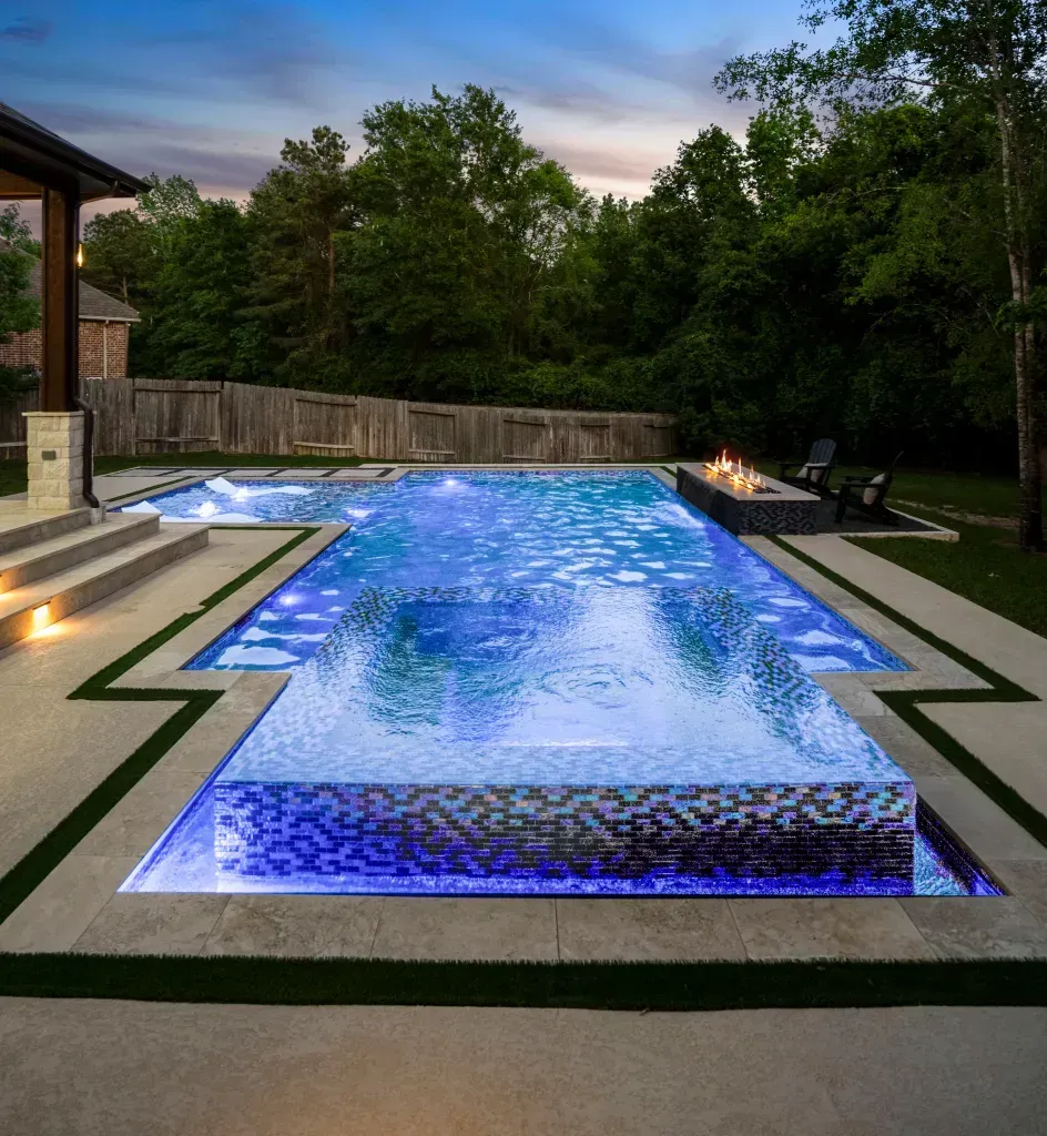 Modern backyard pool with blue-lit water, stone decking, and trees at dusk