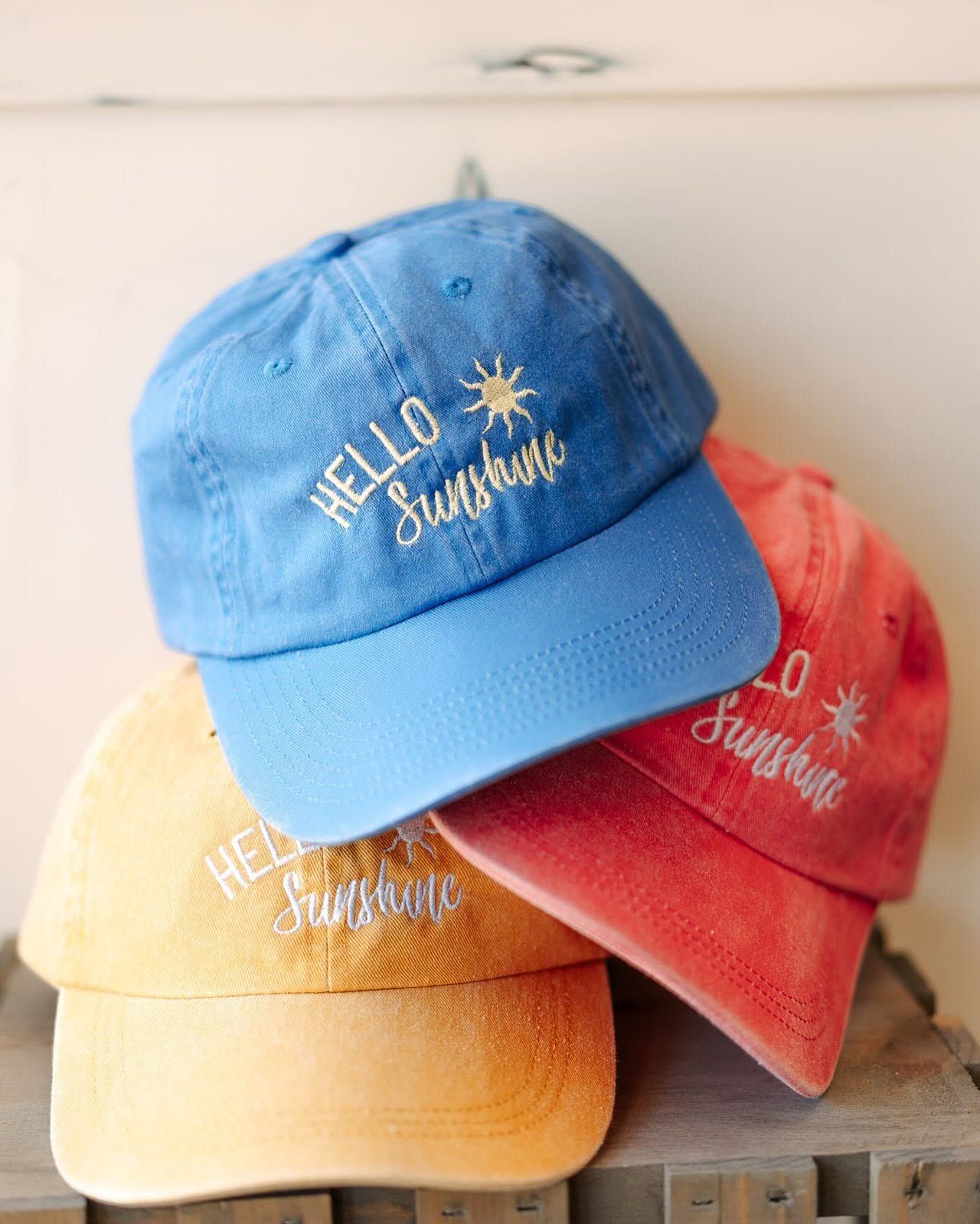 Three stacked “Hello Sunshine” baseball caps in blue, red, and yellow