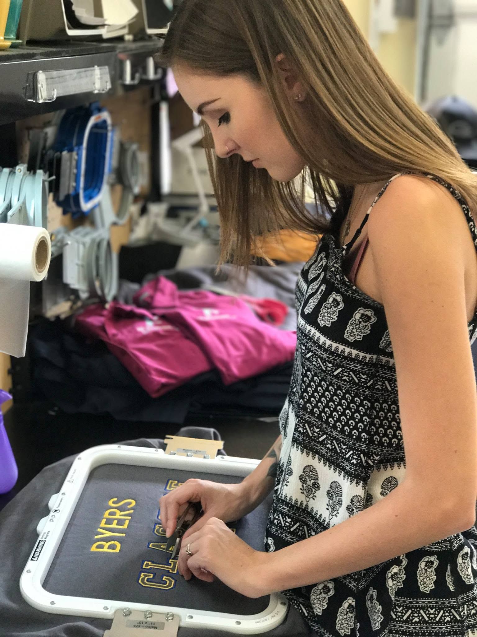 Woman placing gold lettering on a gray shirt at a craft table with pink supplies nearby